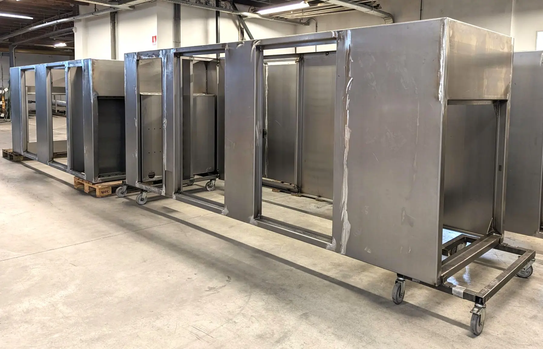 Onafgewerkte roestvrijstalen kastframes met zichtbare lasnaden staan op verrijdbare onderstellen in een industriële werkplaats.