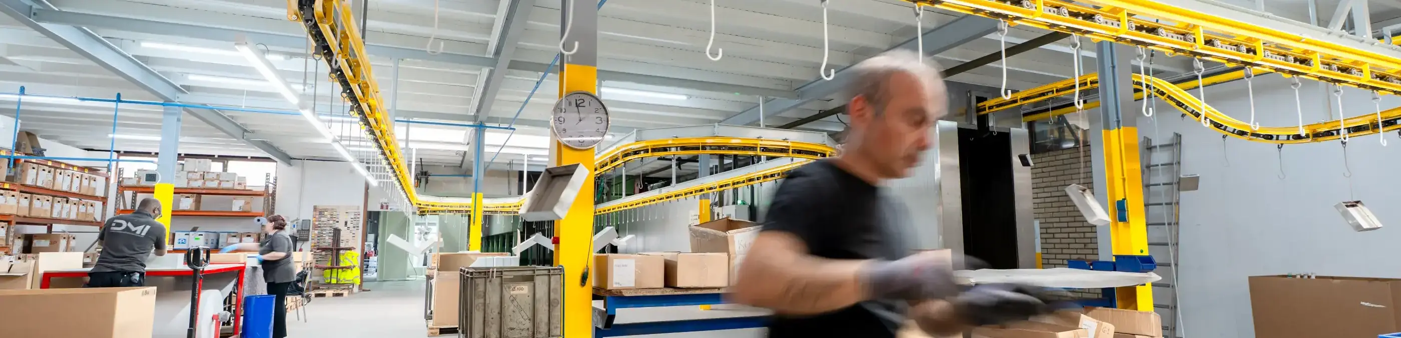 Een man in zwarte kleding werkt met handschoenen aan metalen platen in een hal met gele hangende transportband en dozen.