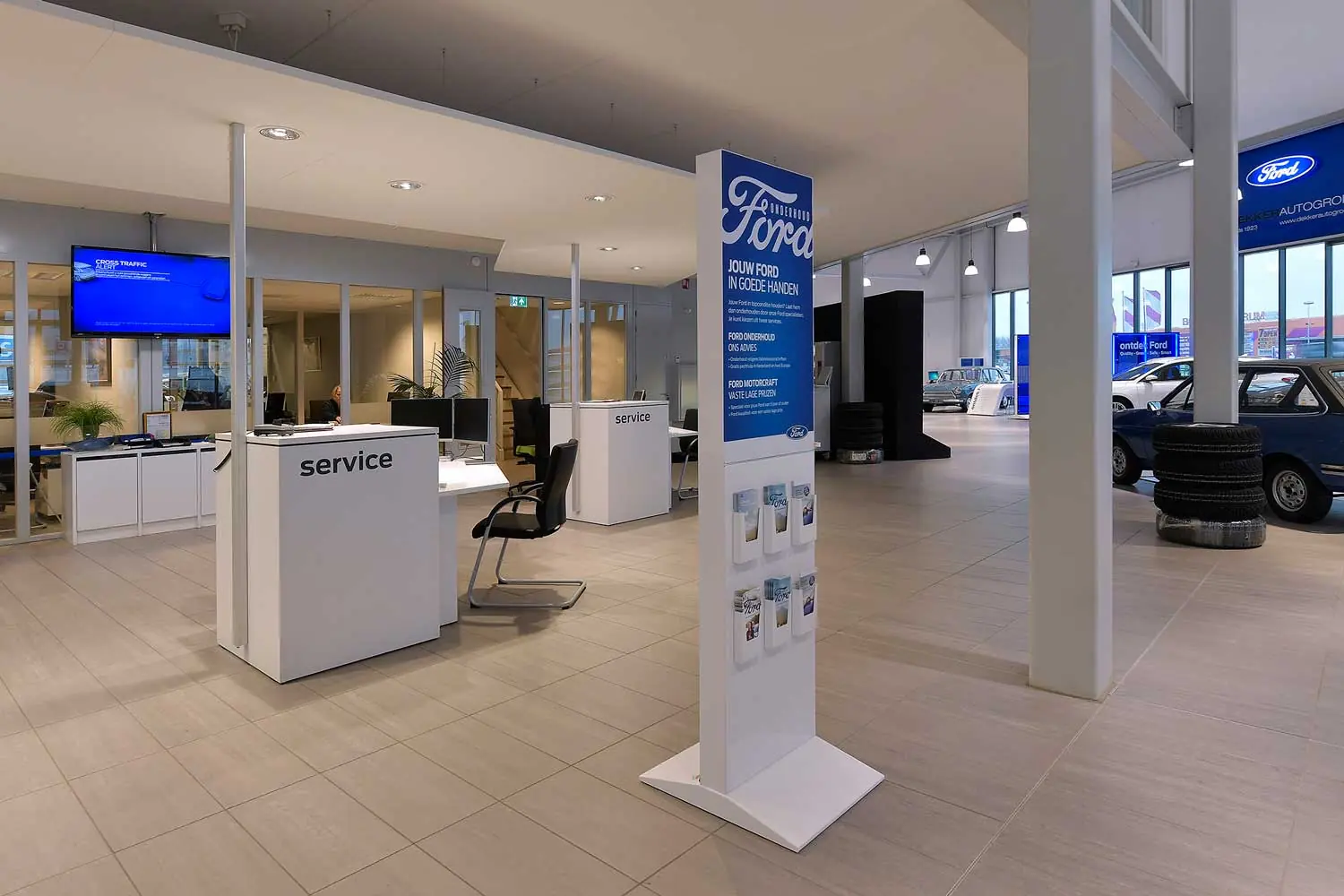 Ruime Ford-showroom met witte servicebalies, een blauw Ford-onderhoudsbord met folders en een geparkeerde auto met banden op de achtergrond.