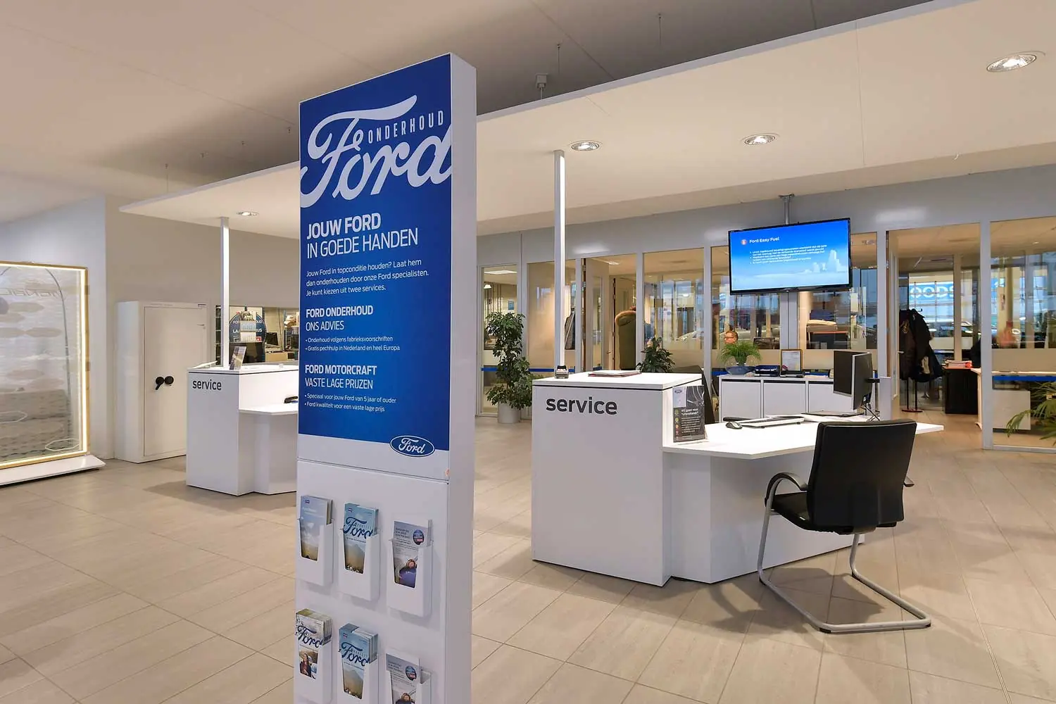 Witte servicebalies met bureaustoel en een blauwe Ford Onderhoud-zuil met folders in een lichte showroom met glazen wanden.