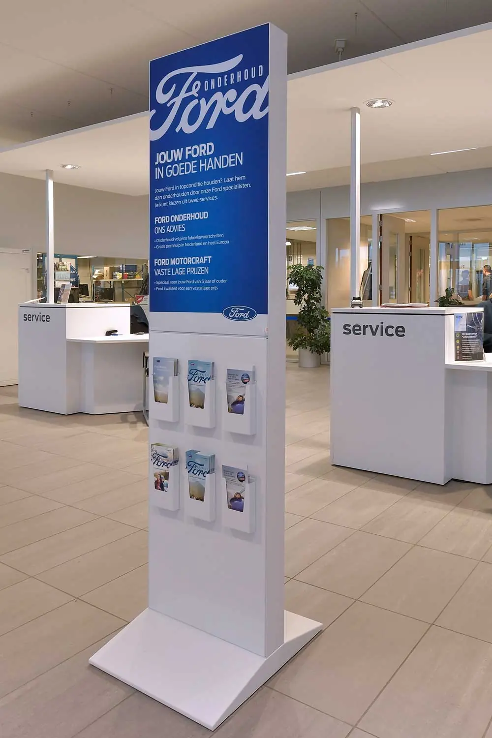 Blauwwitte Ford-zuil met folderhouders in een showroom, met twee servicebalies en een plant op de achtergrond.