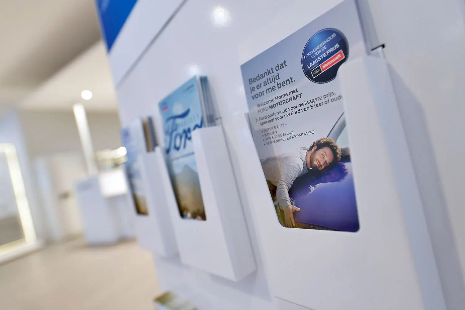 Witte folderhouders aan de wand met Ford-brochures, voorop een folder over Ford Motorcraft onderhoud voor de laagste prijs.