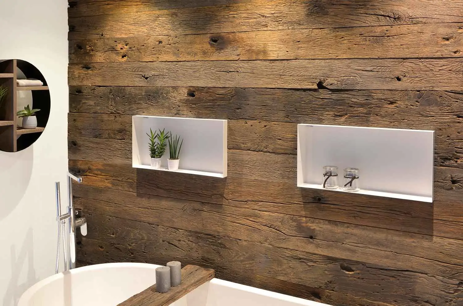 Badkamer met wand van verweerd hout, twee witte nissen met planten en glaasjes, een vrijstaand bad en een chromen kraan.