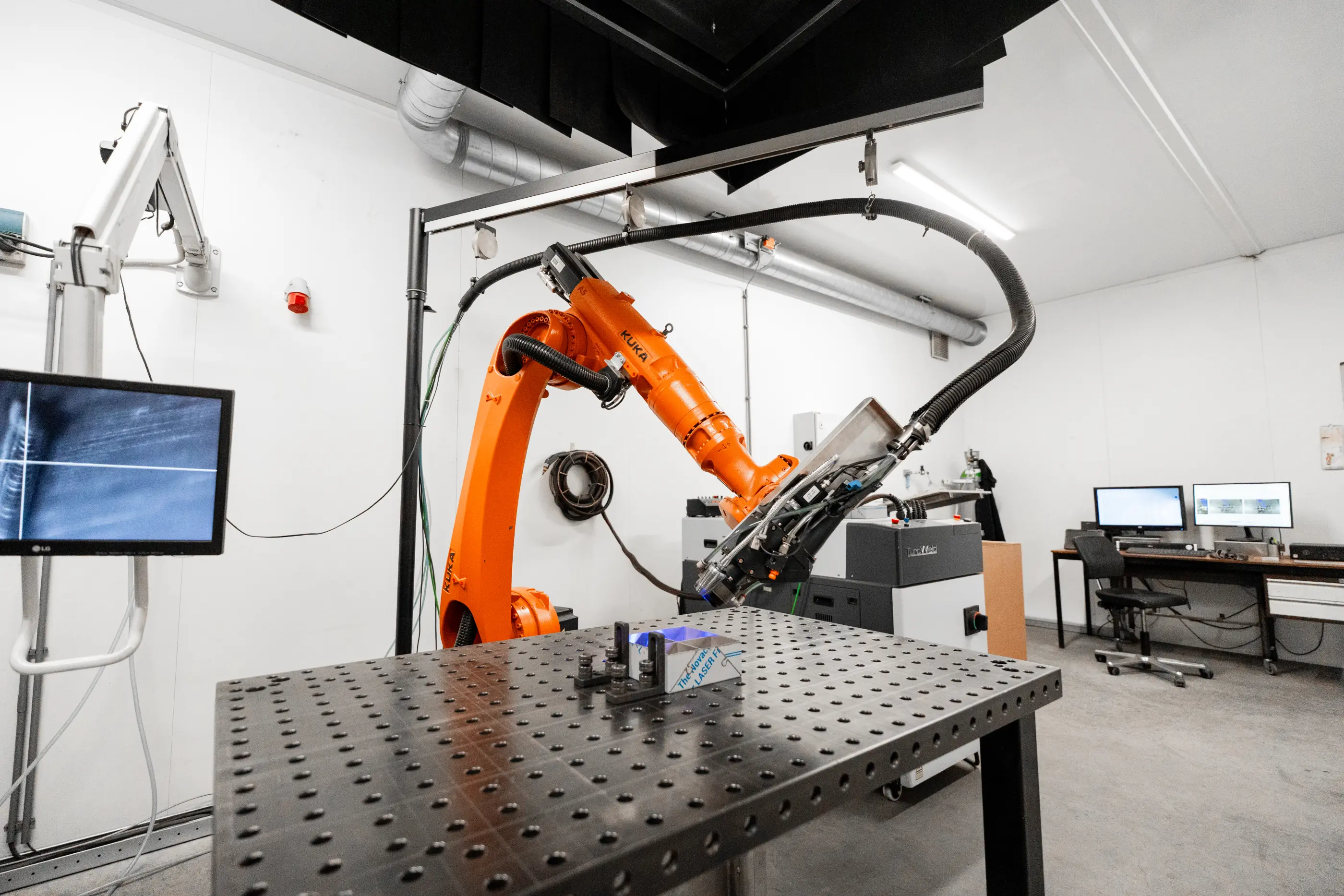 Oranje KUKA-robotarm met laserkop boven een geperforeerde werktafel met ingespannen werkstuk, met monitor en computers in een witte ruimte.