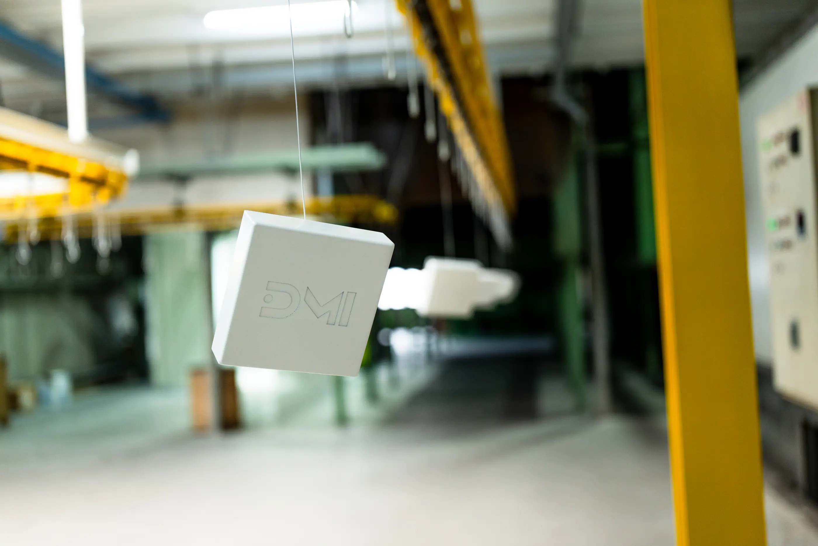 Wit blok met DMI-logo hangt aan een draad in een industriële hal met gele railconstructies en een bedieningskast op de achtergrond.