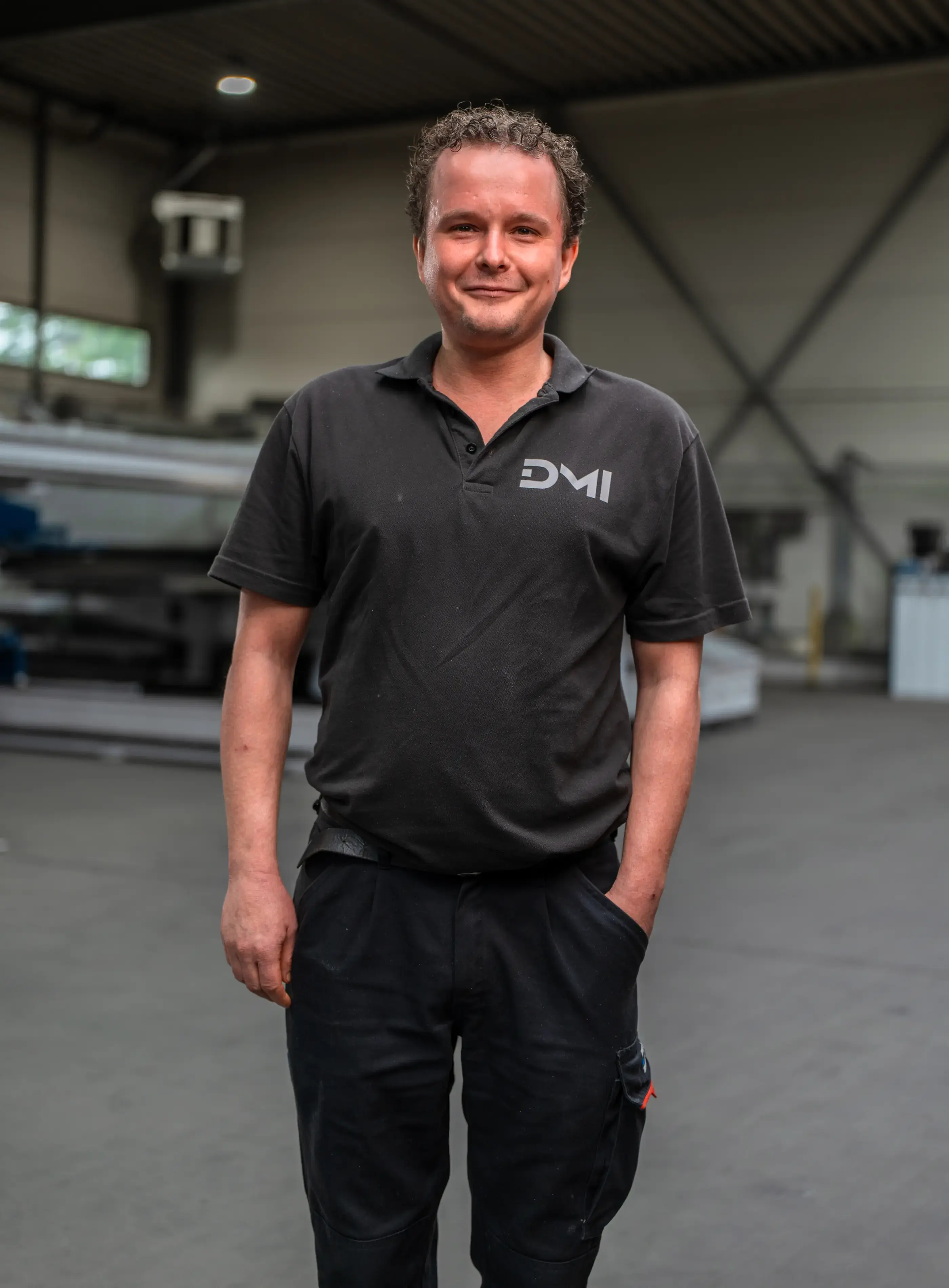 Glimlachende man met krullend haar in zwart poloshirt met DMI-logo staat met een hand in zijn zak in een industriële werkhal.