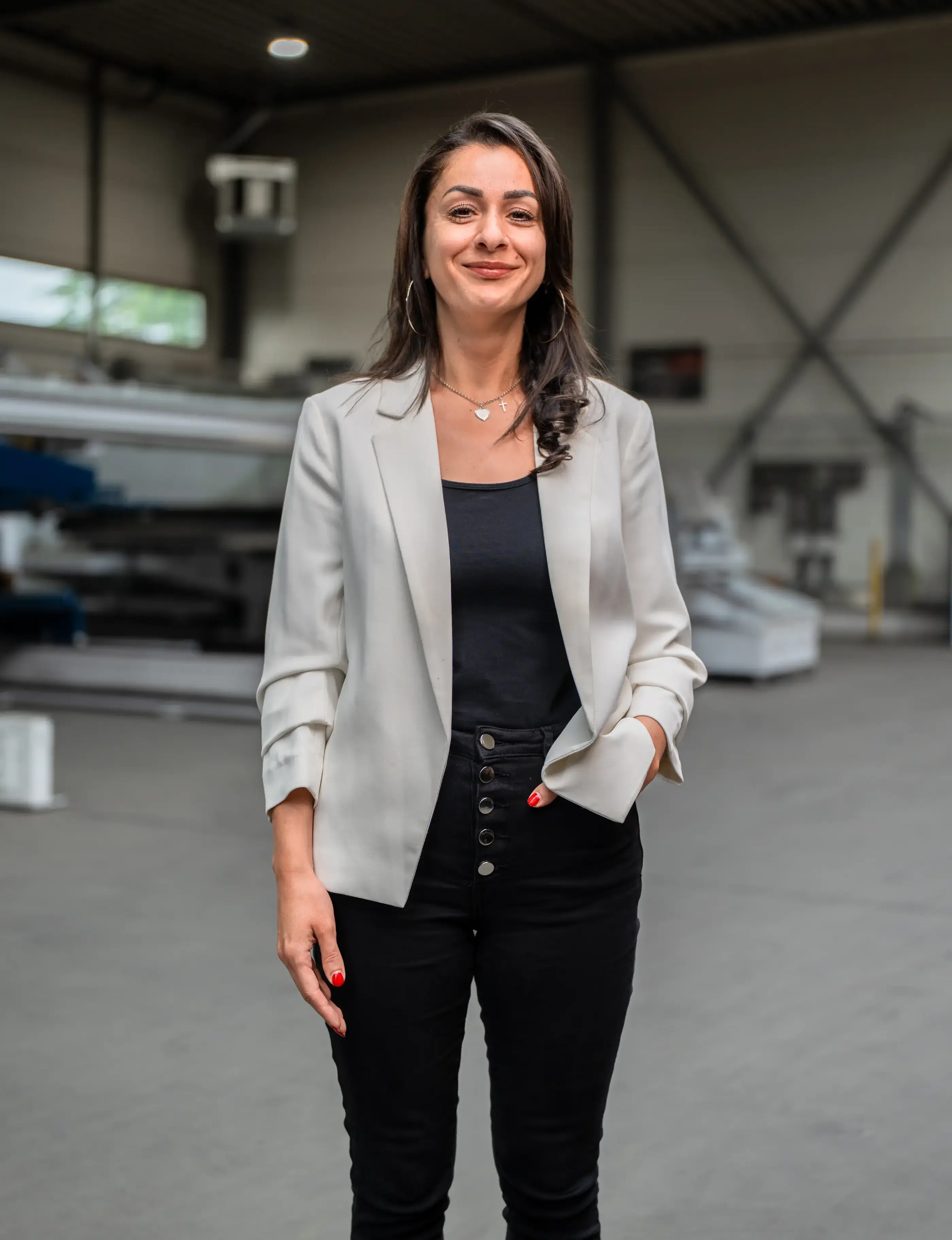 Glimlachende vrouw in lichte blazer en zwarte broek staat met een hand in de zak in een grote industriële hal.