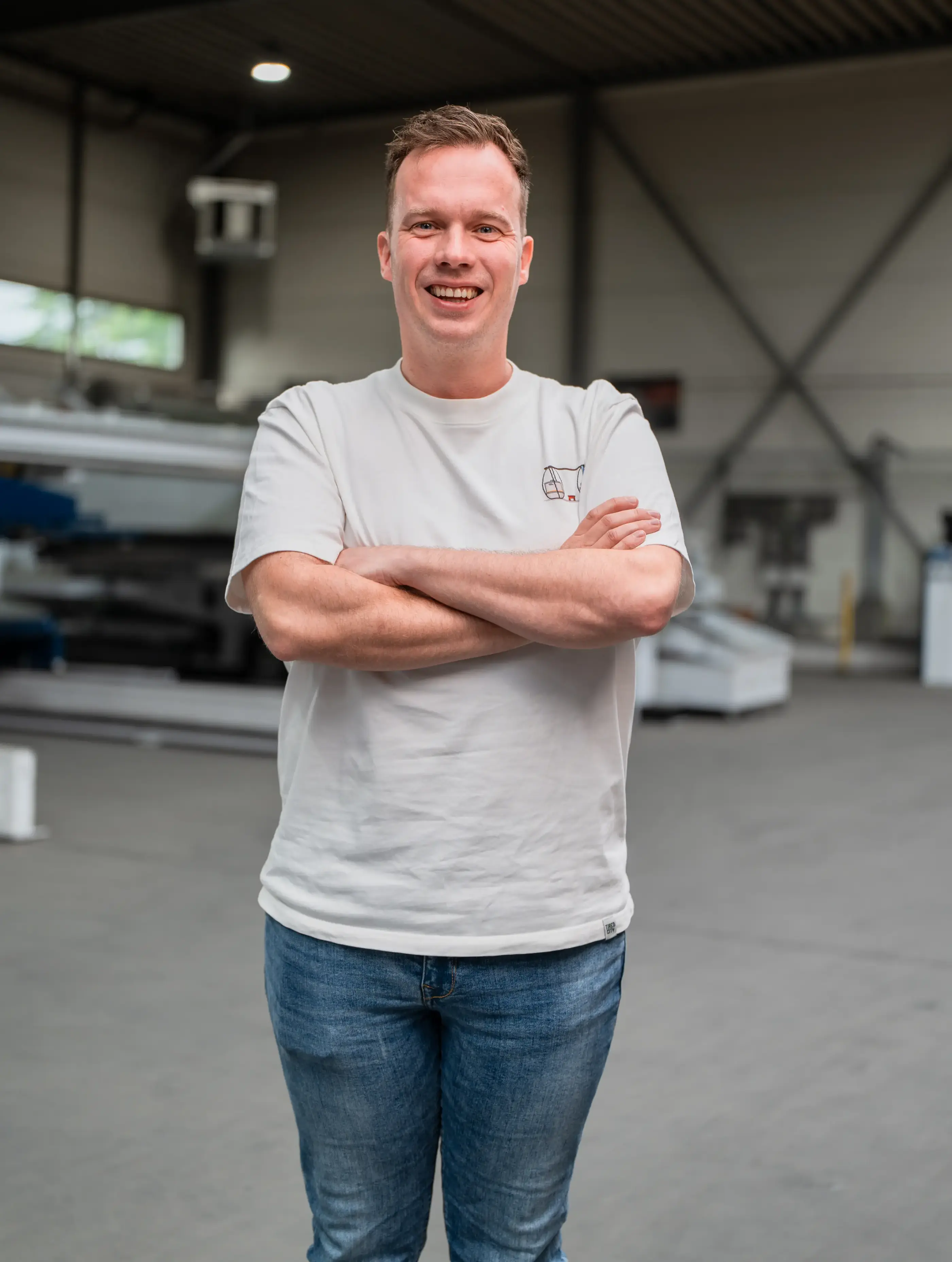 Lachende man in wit t-shirt en spijkerbroek staat met over elkaar geslagen armen in een ruime industriële hal met machines.