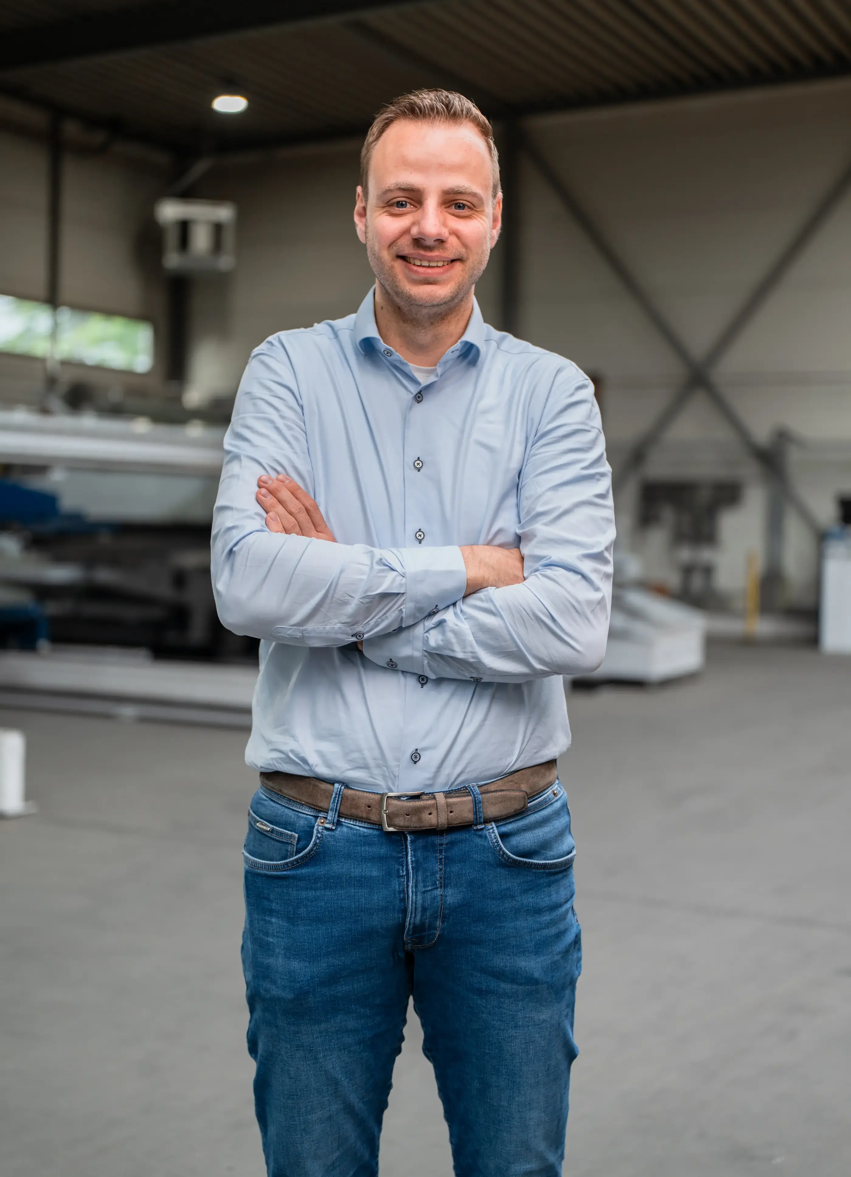 Glimlachende man in lichtblauw overhemd en spijkerbroek staat met gekruiste armen in een industriële werkhal.