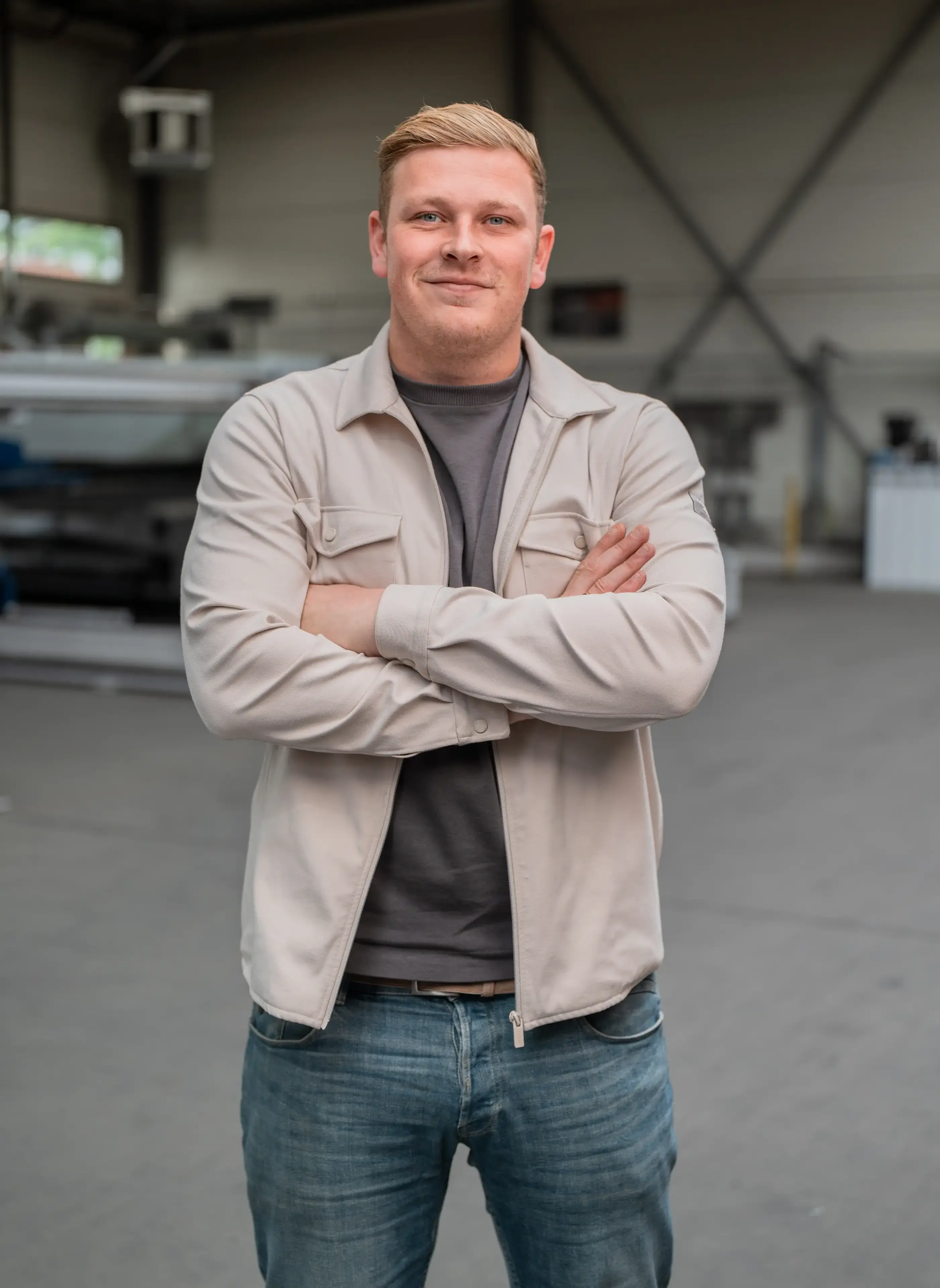 Man met gekruiste armen, beige overshirt en spijkerbroek, glimlachend in een bedrijfshal met machines op de achtergrond.
