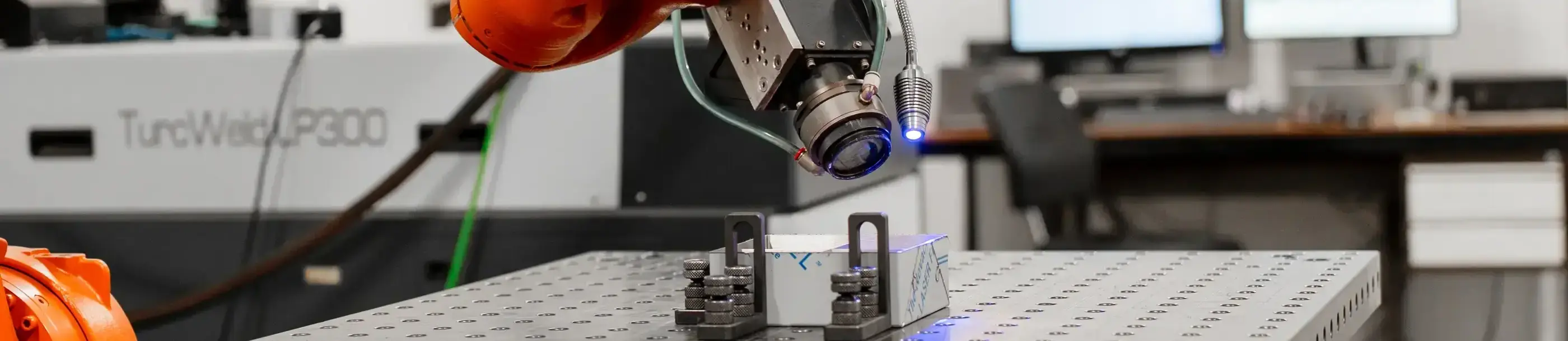 Oranje KUKA-robotarm met laserkop richt blauw licht op een ingespannen werkstuk op een geperforeerde lastafel in een werkplaats.