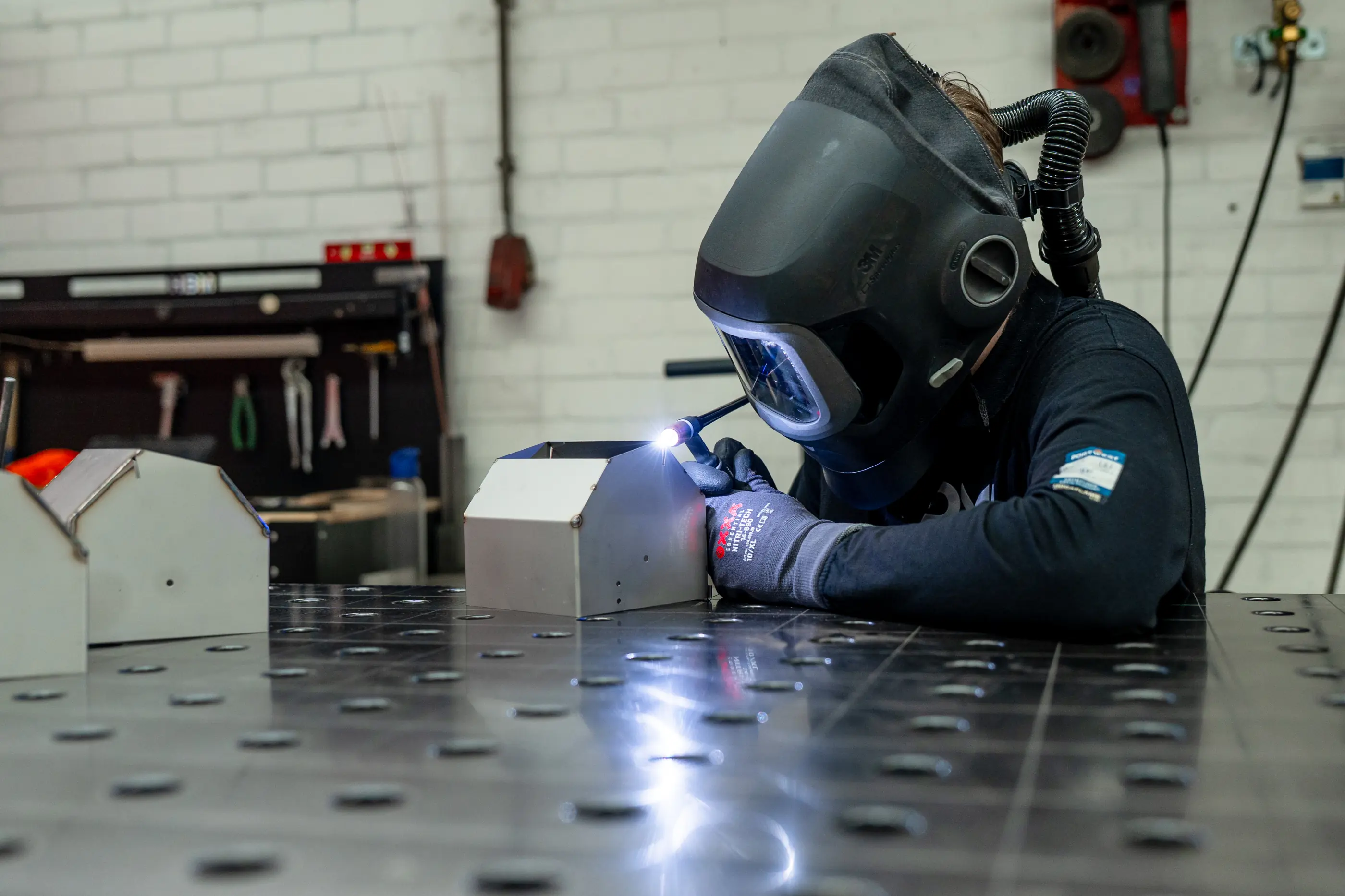 Lasser met lashelm en beschermende handschoenen last aan een metalen werkstuk op een lastafel in een werkplaats.