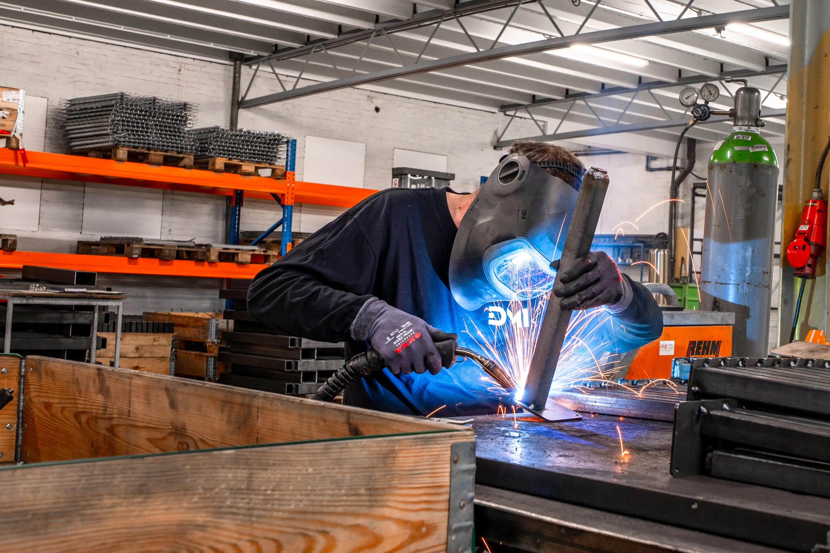 Een lasser met helm en handschoenen last een metalen buis op een plaat, terwijl er vonken rondvliegen in een werkplaats.