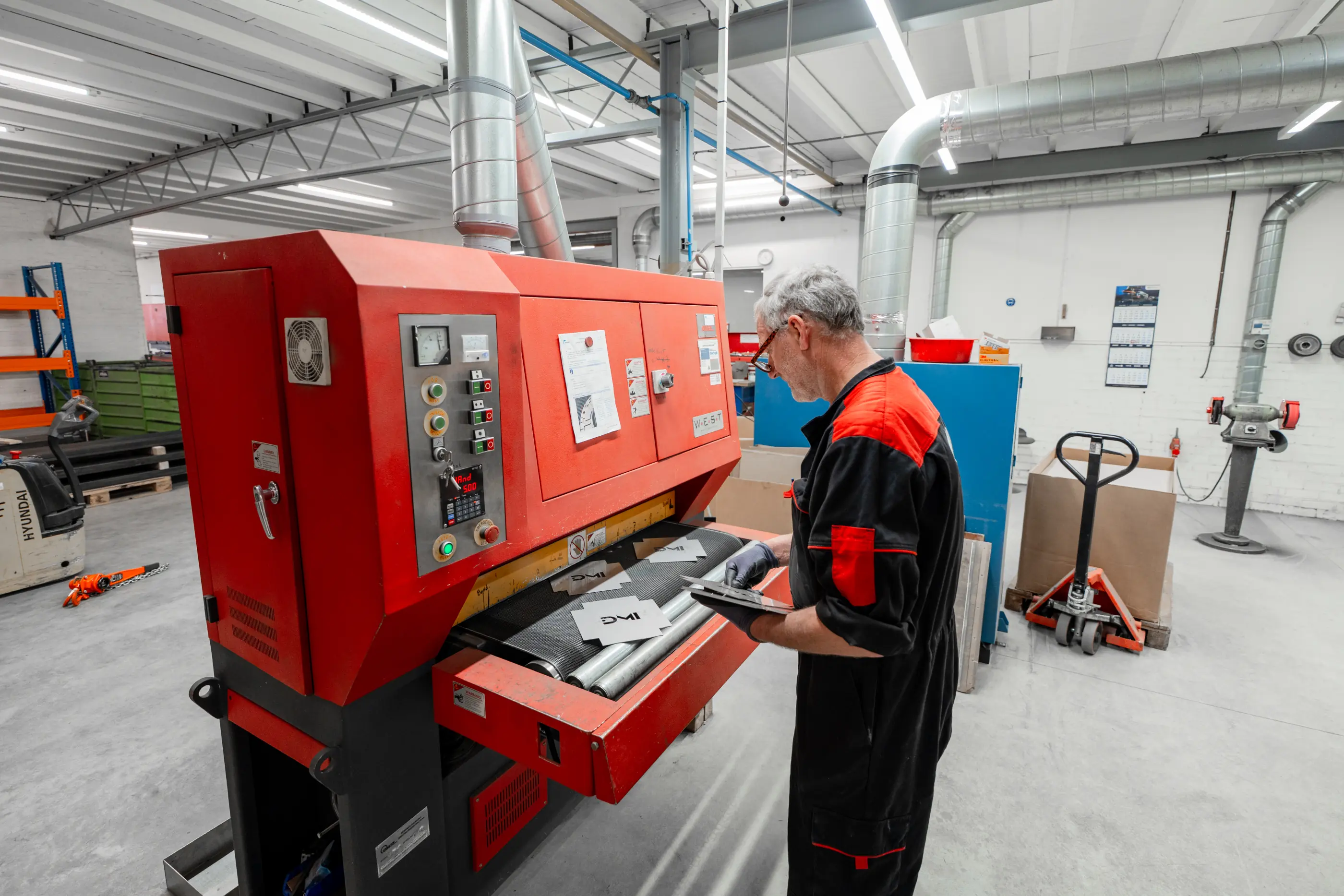 Man in zwart-rode overall bedient een rode bandschuurmachine met metalen plaatjes in een werkplaats met afzuigbuizen.