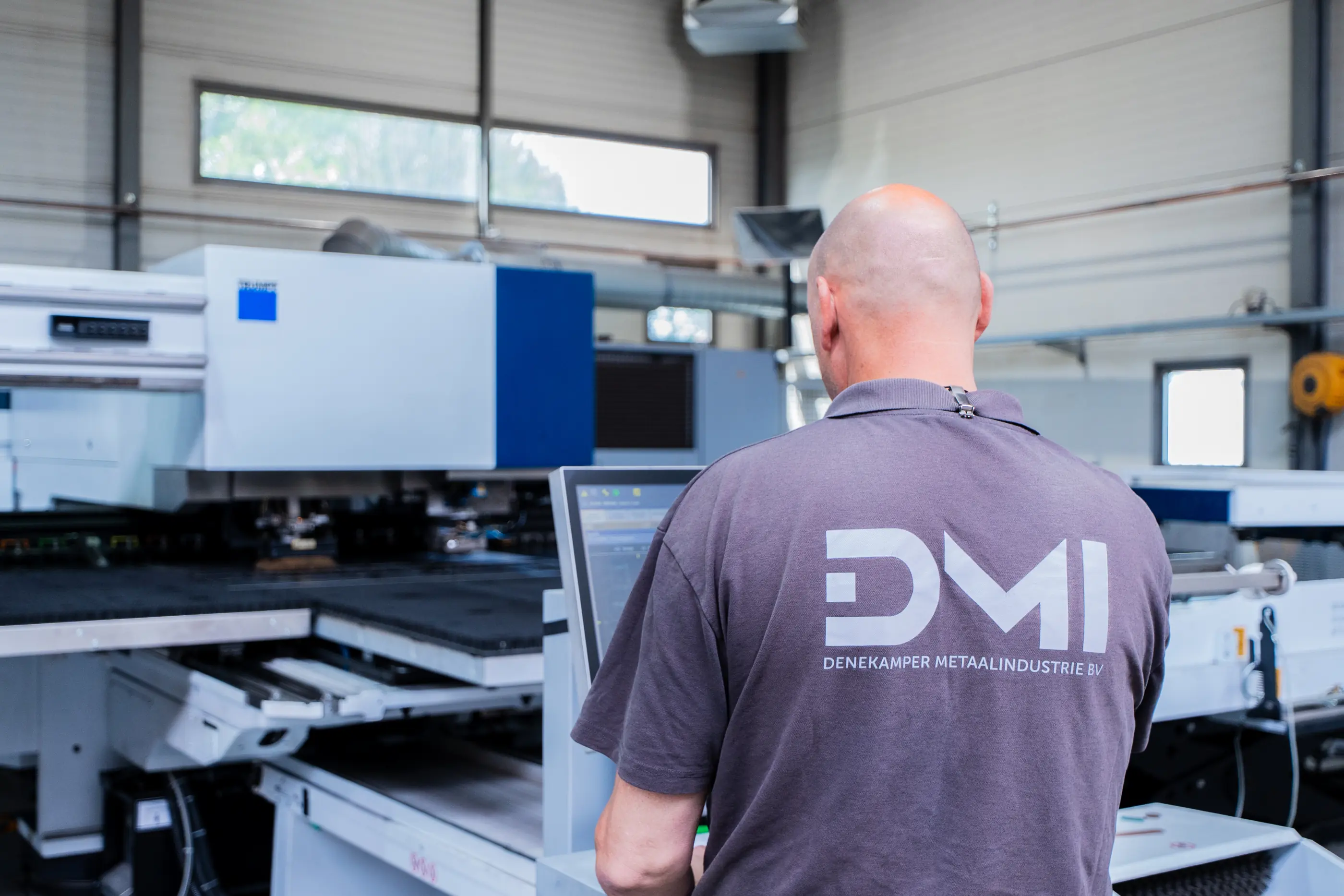 Man in grijs DMI-shirt van achteren bedient het scherm van een ponsmachine in een werkhal van Denekamper Metaalindustrie.