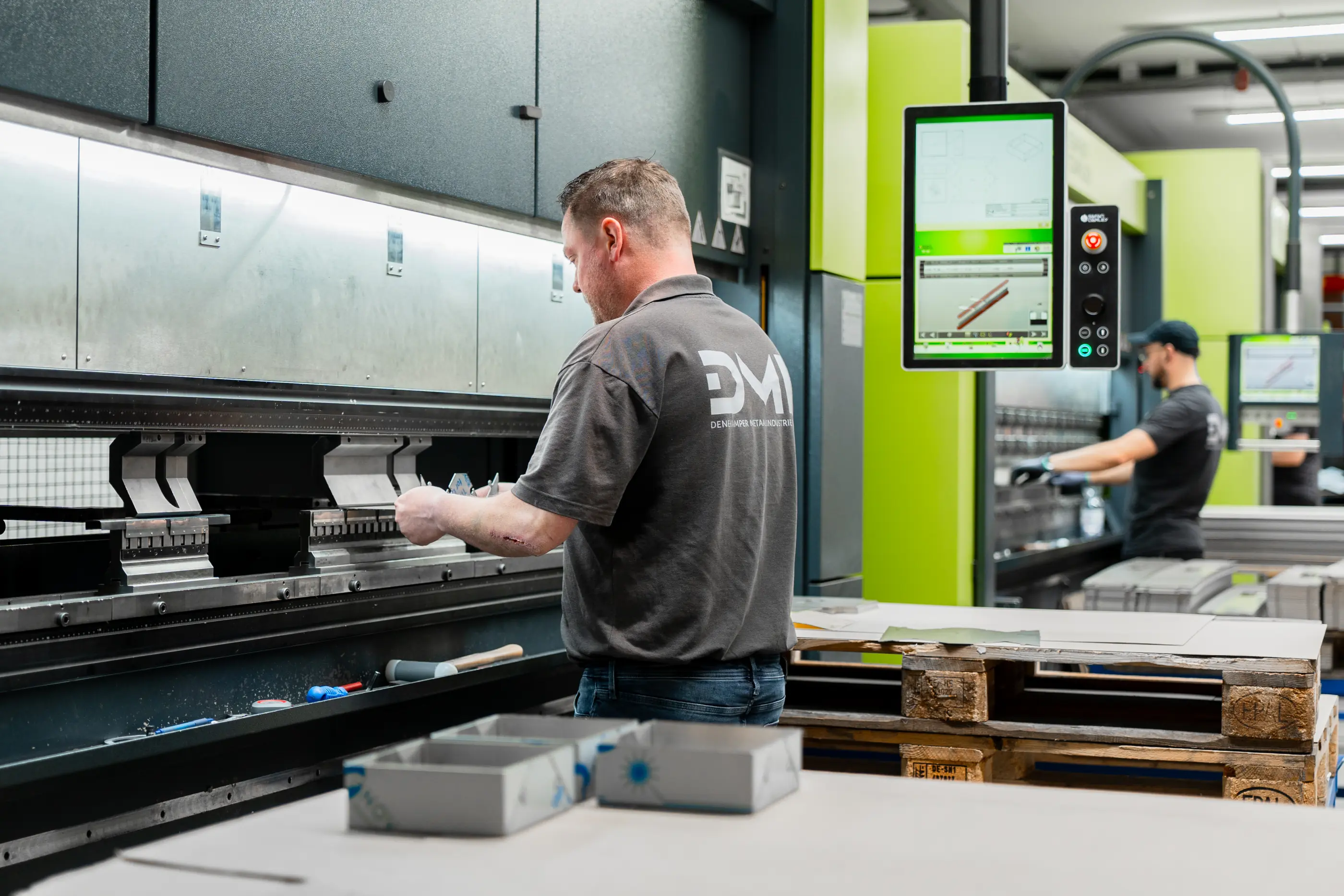 Medewerker in grijs shirt bedient een kantbank in een werkplaats; op de achtergrond een collega en een bedieningsscherm.