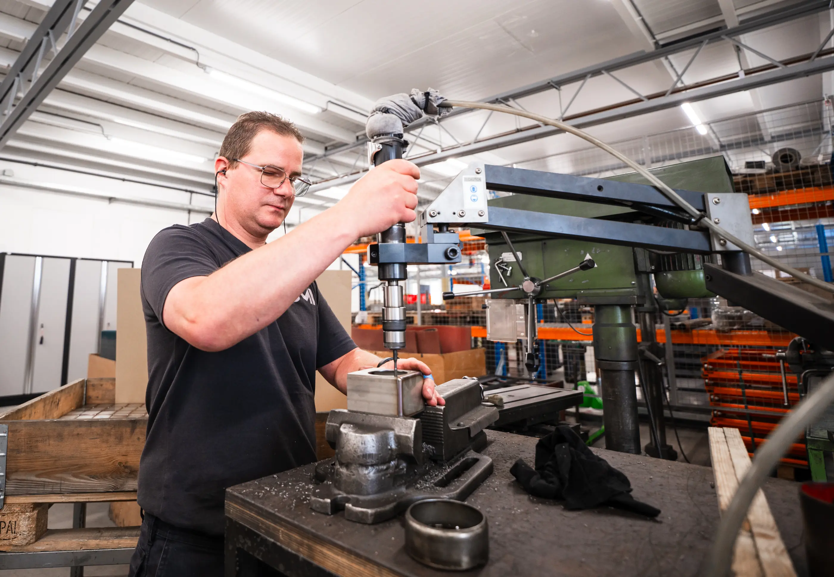Man met bril tapt een metalen werkstuk dat in een bankschroef zit, met een taparm aan een kolomboormachine in een werkplaats.