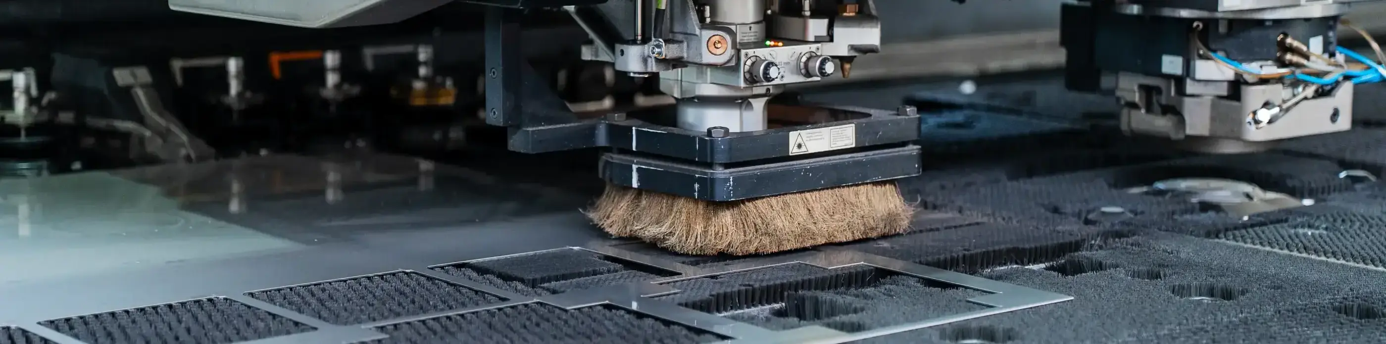 Close-up van een ponsmachine met borstelkop boven een geperforeerde metalen plaat op een werktafel met zwarte borstelpinnen.