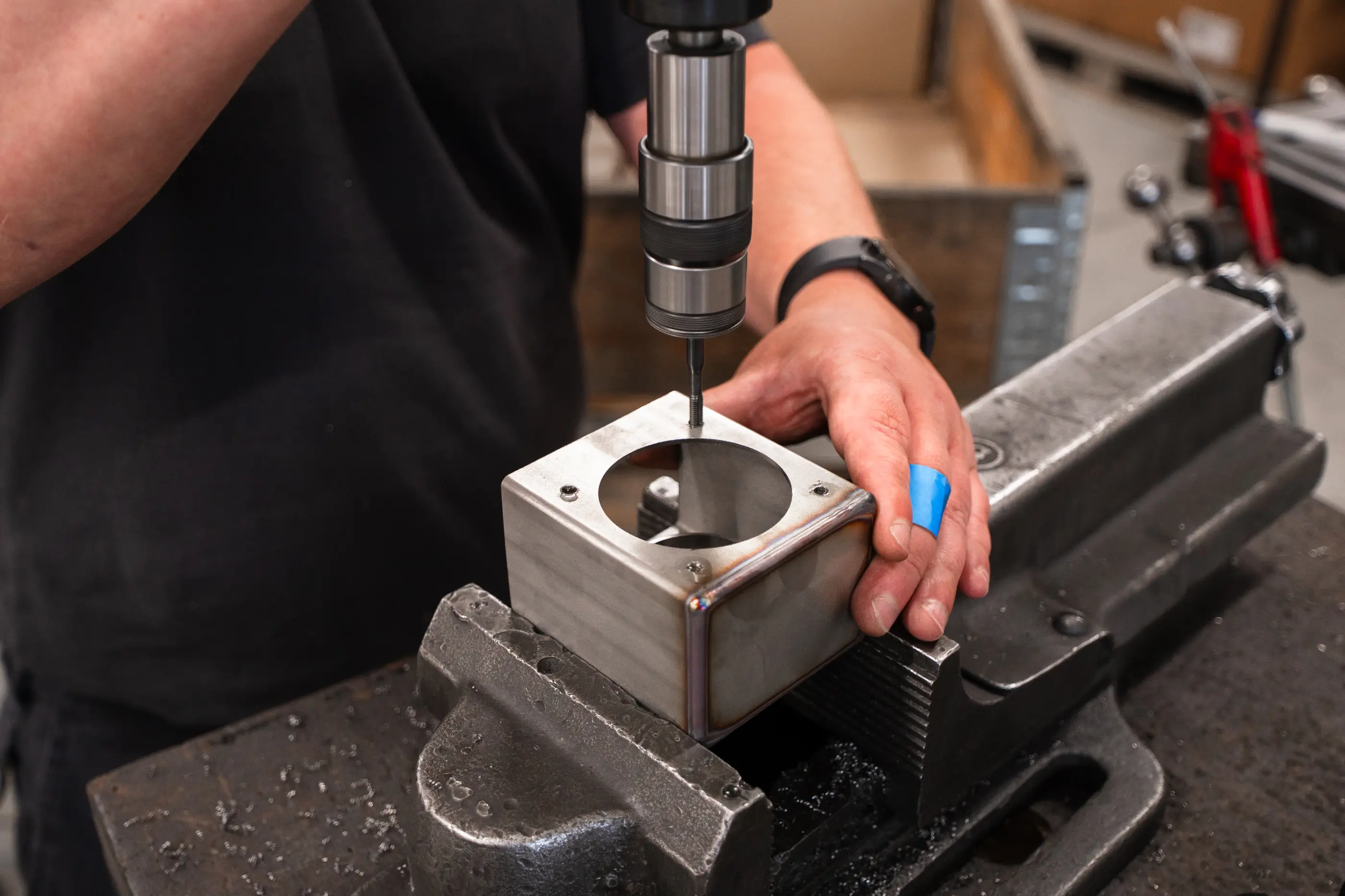 Een hand houdt een metalen behuizing met ronde uitsparing vast in een bankschroef terwijl een boor of tapkop erboven staat.