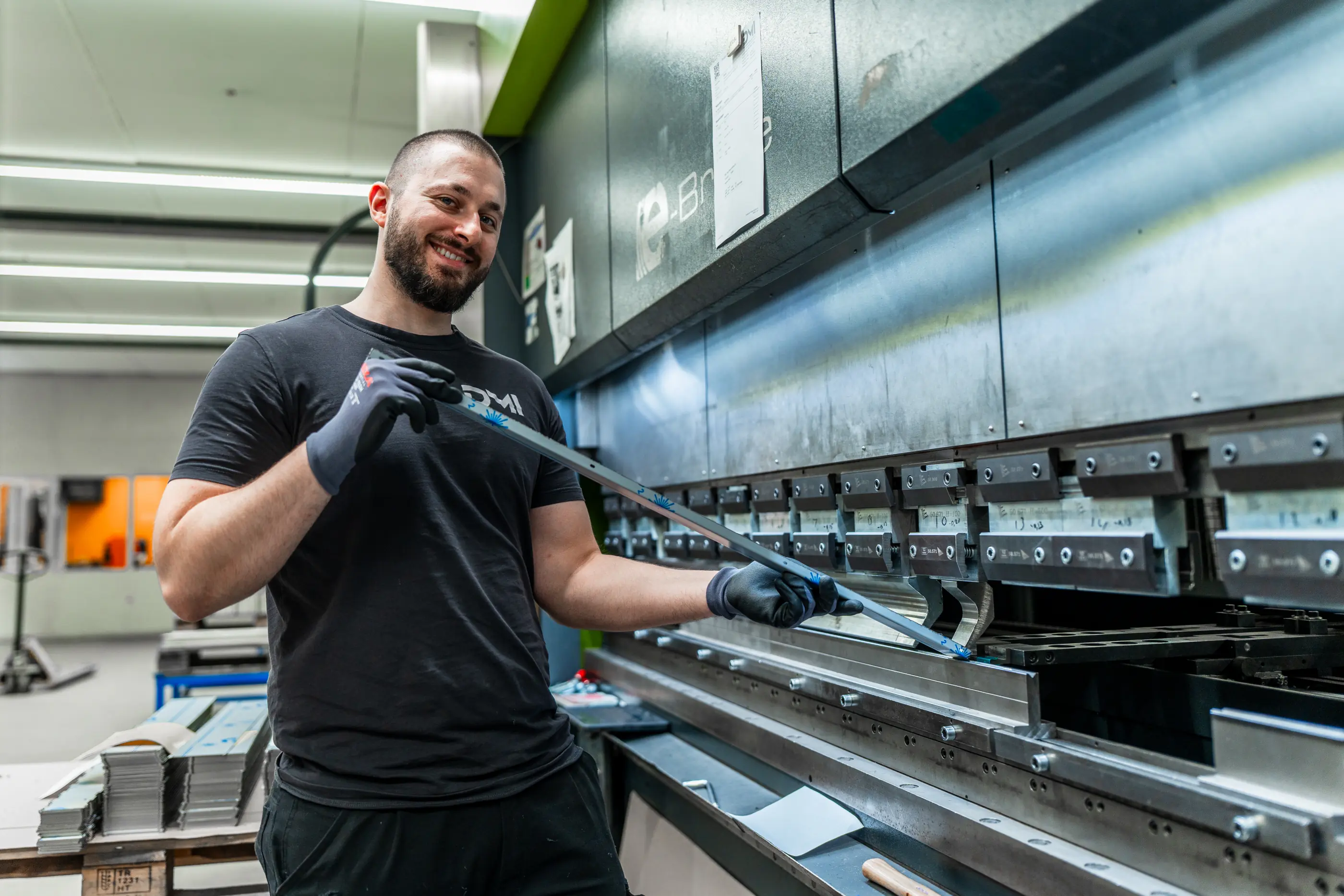 Lachende man met handschoenen houdt een metalen strip vast bij een kantbank in een werkplaats, met stapels plaatwerk achter hem.