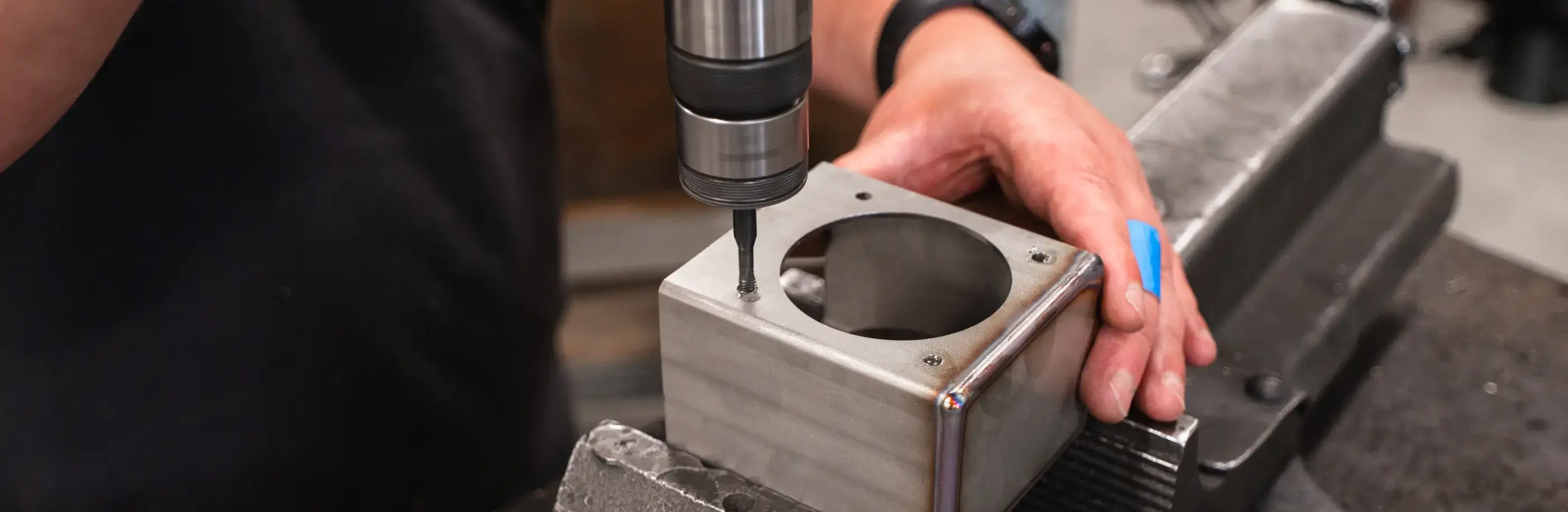 Een man houdt een metalen kastje met groot rond gat in een bankschroef terwijl een tapmachine een klein gat bewerkt.