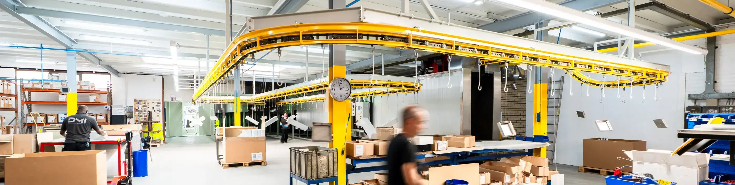 Werkplaats met gele hangende transportbaan, kartonnen dozen op pallets en medewerkers aan het werk in een lichte industriële hal.
