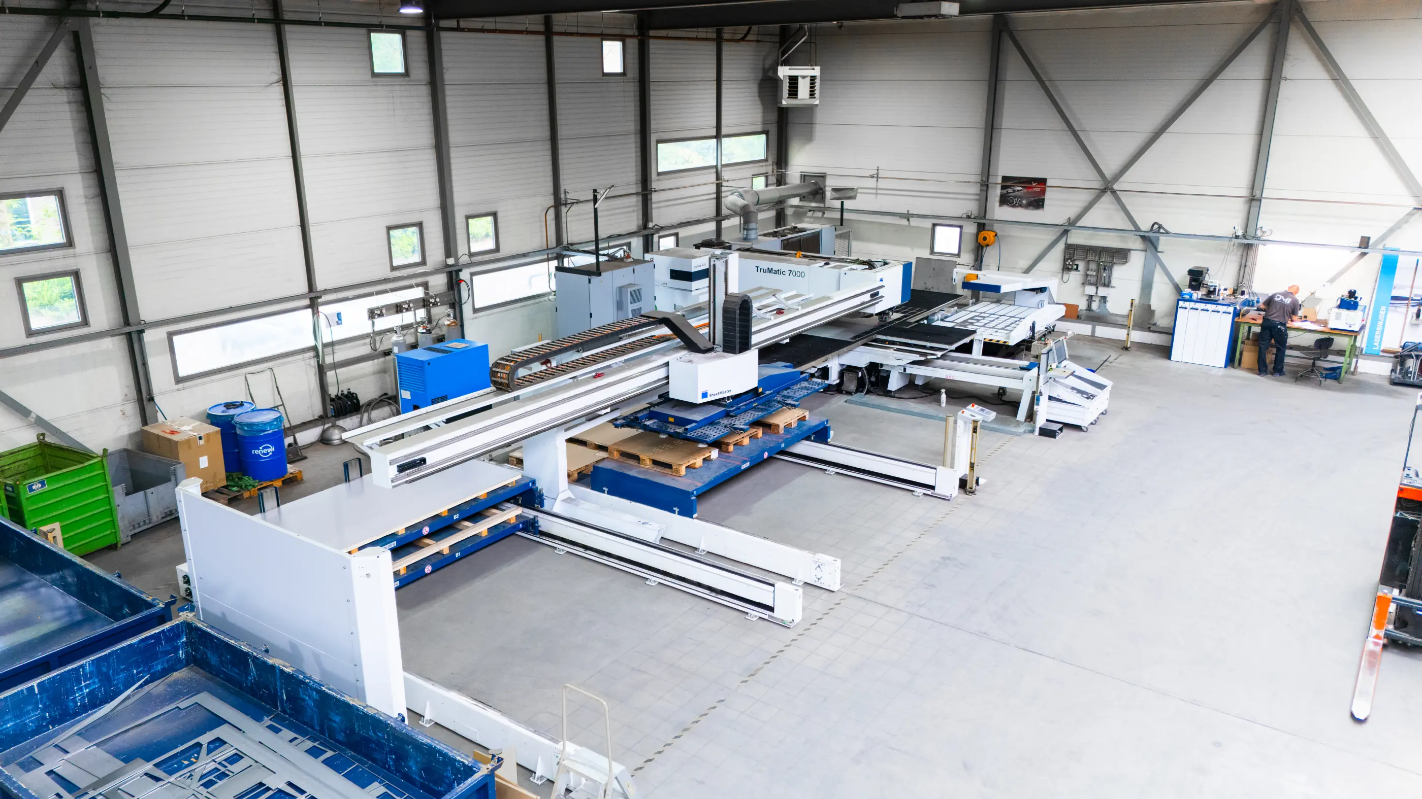 Overzicht van een industriële hal met een TruMatic 7000 plaatbewerkingsmachine, laadsysteem, pallets en een medewerker aan een werkbank.