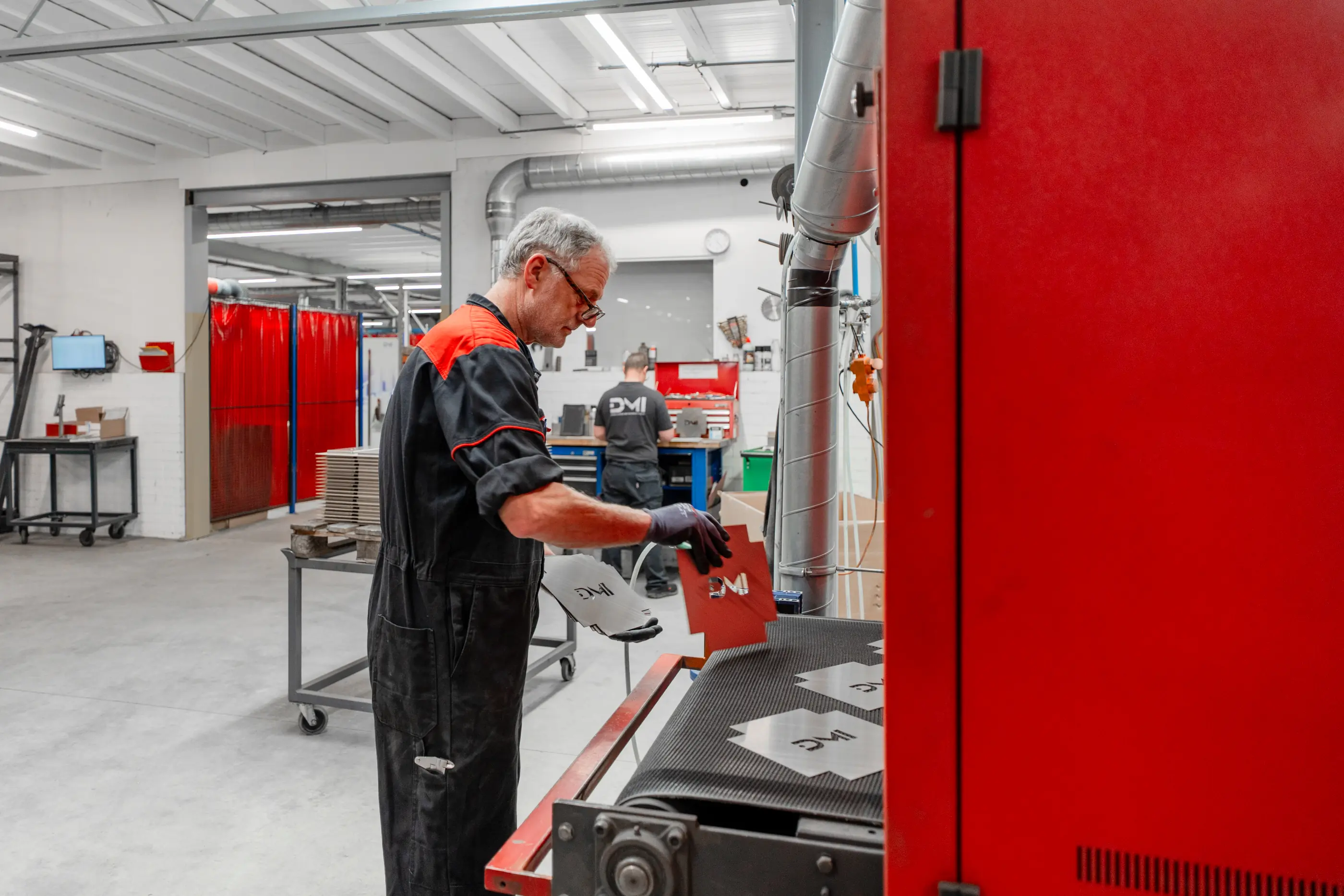 Een man in werkoverall legt een metalen plaatdeel op de transportband van een ontbraammachine in een werkplaats.