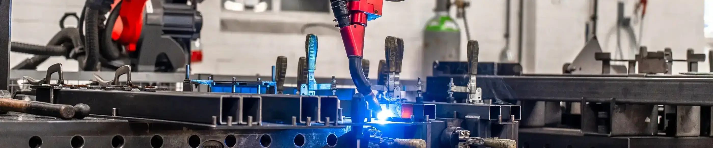 Een rode lasrobot last stalen profielen die op een geperforeerde werktafel met spanklemmen zijn vastgezet in een werkplaats.