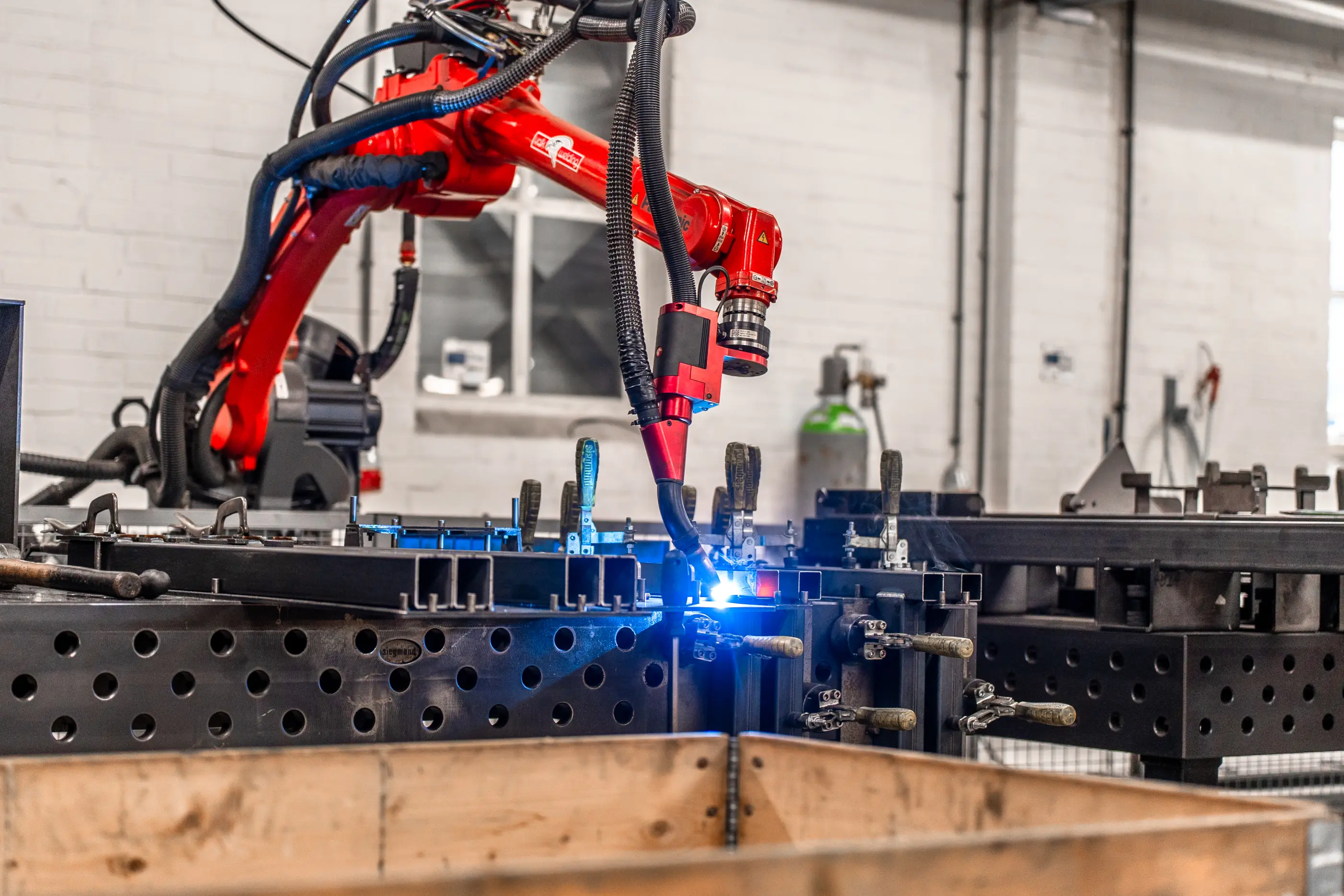 Rode lasrobot last stalen profielen die op een geperforeerde werktafel met spanklemmen zijn vastgezet, met blauwe lasvonk.