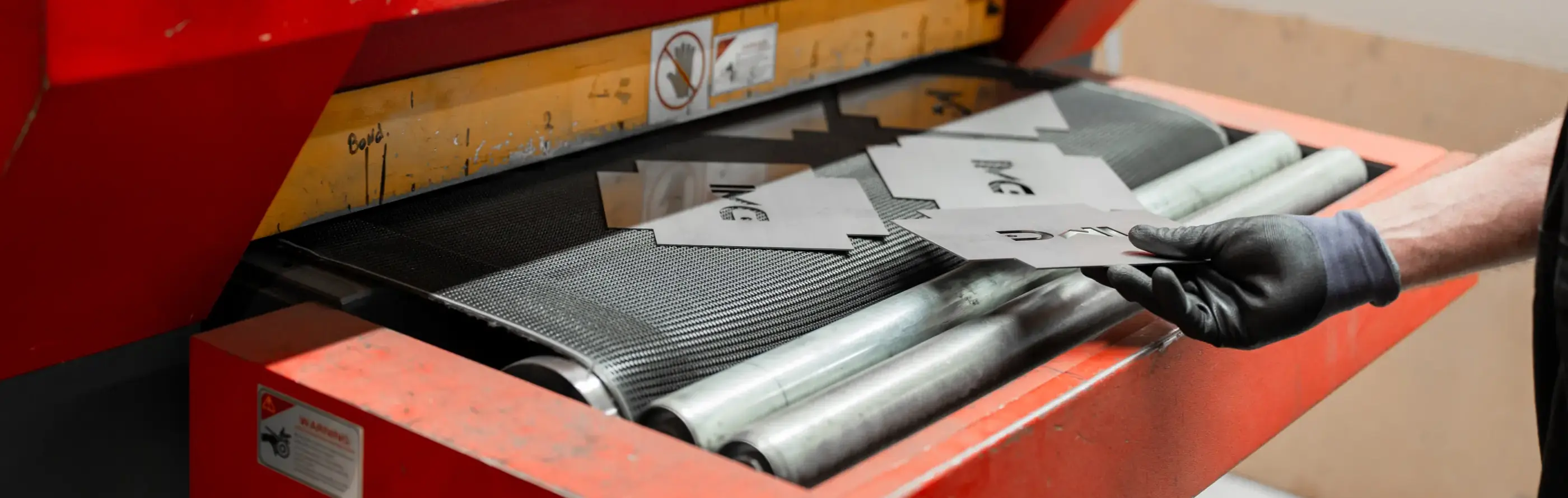 Een hand in zwarte handschoen legt uitgesneden metalen plaatdelen op de transportband van een rode ontbraammachine.