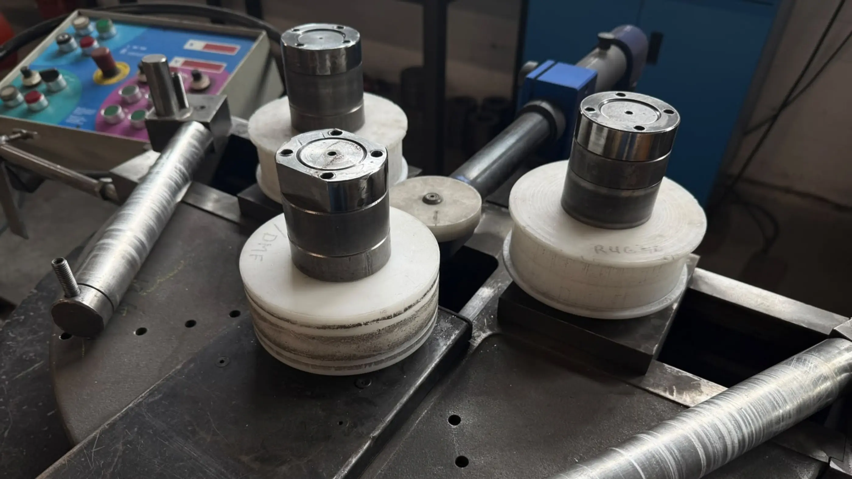 Close-up van een walsmachine met drie witte kunststof rollen op stalen assen, met een bedieningspaneel en metalen buizen op de achtergrond.