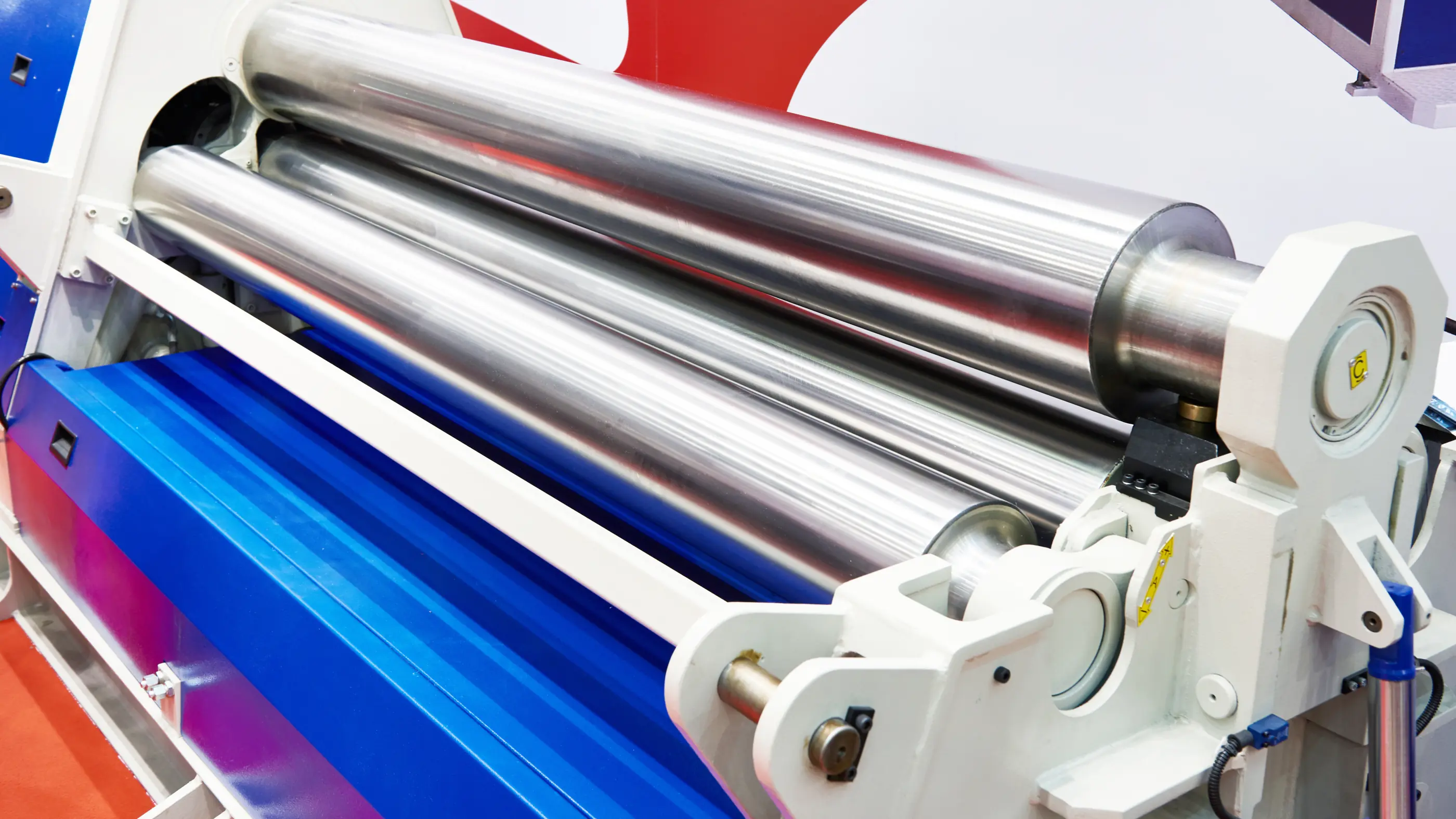 Industriële walsmachine met drie glanzende metalen rollen in een wit en blauw frame, met gele waarschuwingsstickers.