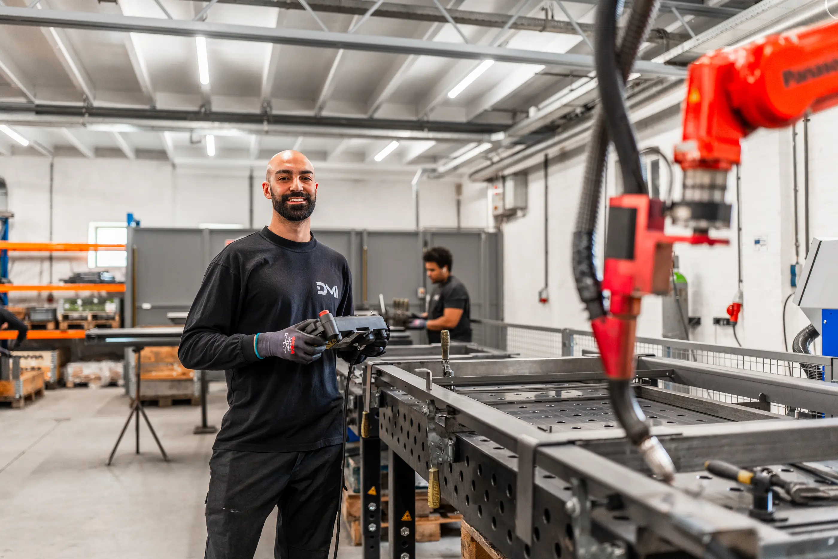Lachende medewerker in zwart shirt houdt een besturingskastje vast naast een rode lasrobot boven een metalen werktafel in een werkhal.