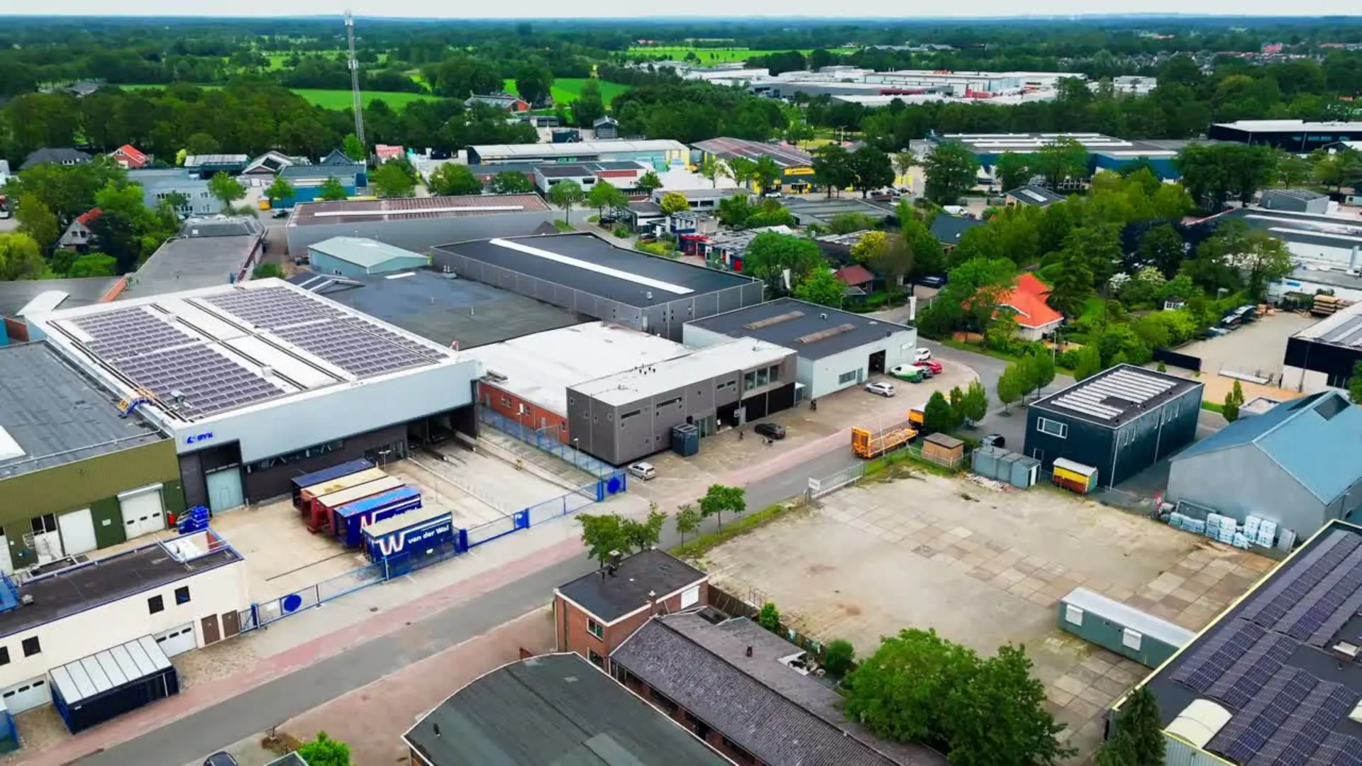 Luchtbeeld van een bedrijventerrein met loodsen, zonnepanelen op daken, geparkeerde auto's, containers en veel groen eromheen.