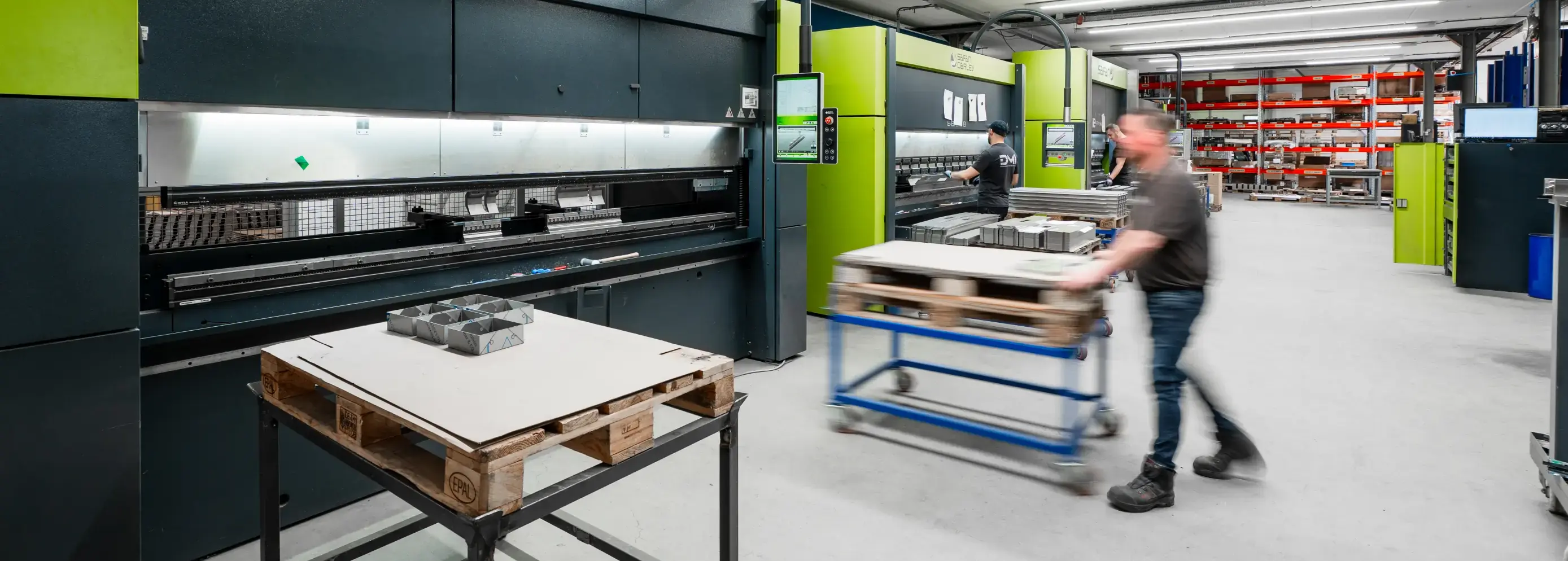 Werknemer duwt een rolwagen door een werkplaats met groene Safan Darley E-Brake kantbanken en stellingen op de achtergrond.