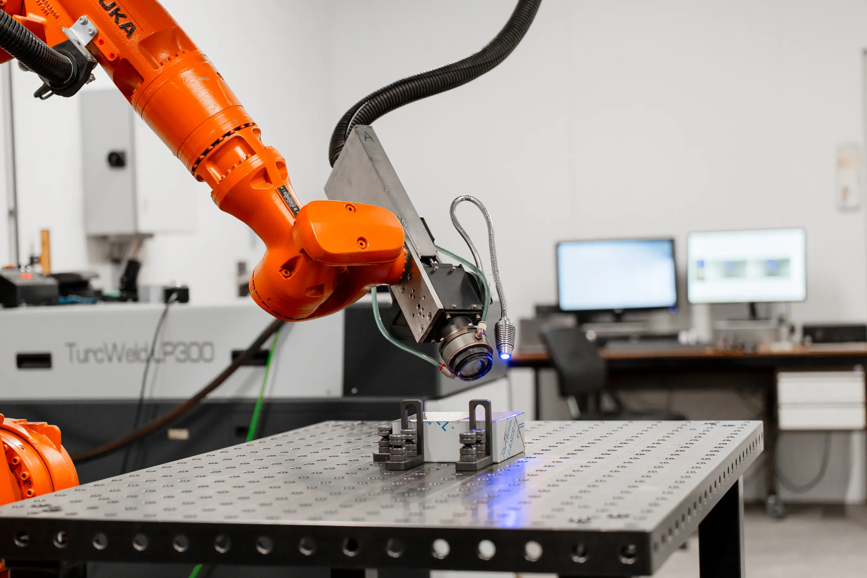 Oranje KUKA-robotarm met meetkop boven een werkstuk op een perforeerde werktafel, met computerschermen op de achtergrond.