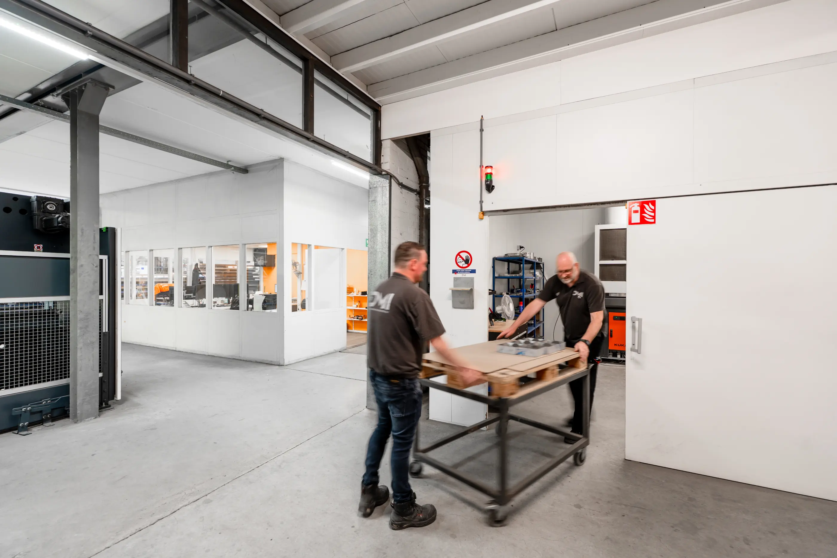 Twee mannen in donkere werkshirts duwen een karretje met een pallet door een industriële werkhal met kantoorruimte op de achtergrond.