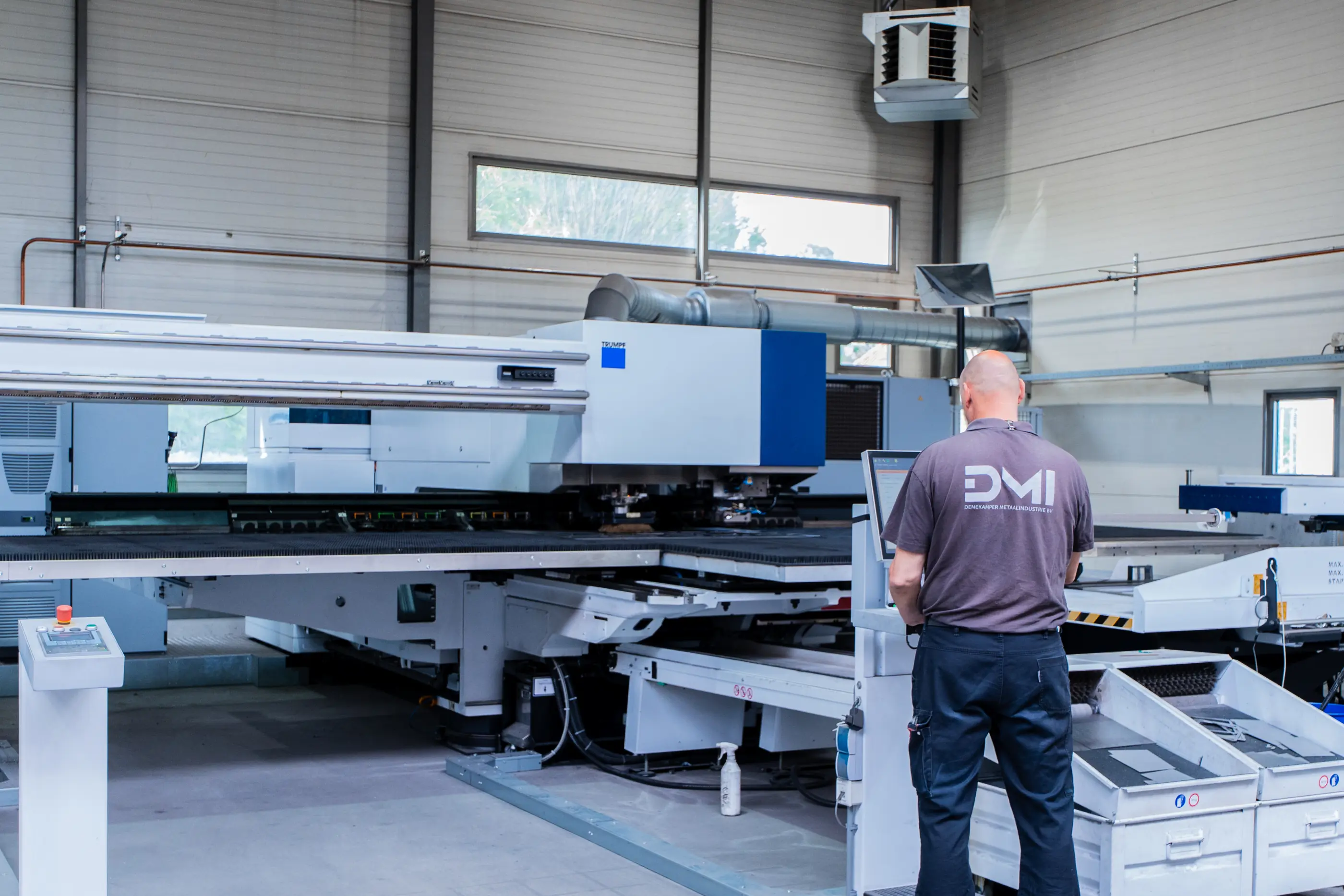 Medewerker in grijs DMI-shirt bedient een grote blauw-witte plaatbewerkingsmachine in een industriële productiehal.