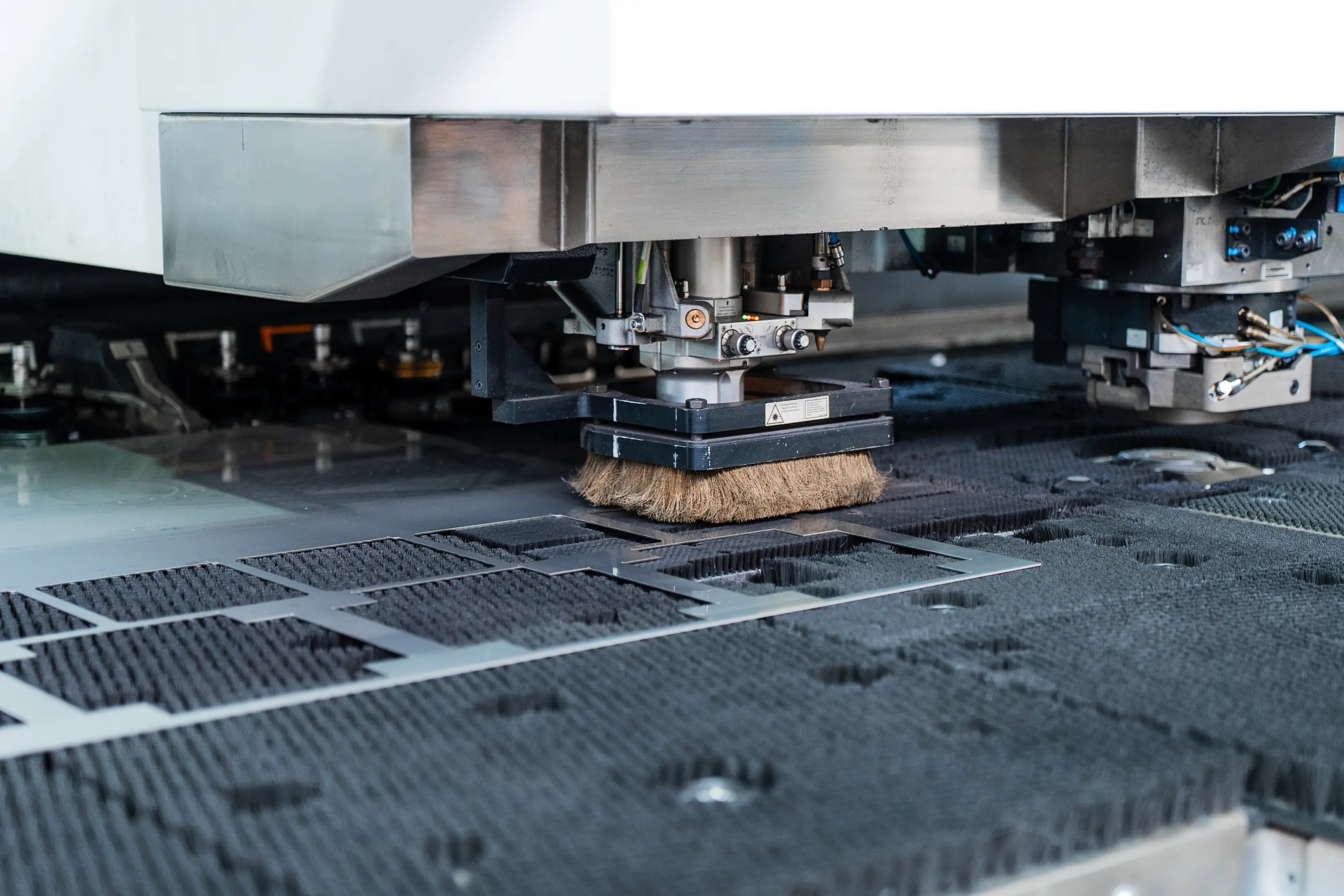 Close-up van een ponsmachine met borstelkop boven een metalen plaat met uitgesneden patronen op een borstelwerktafel.