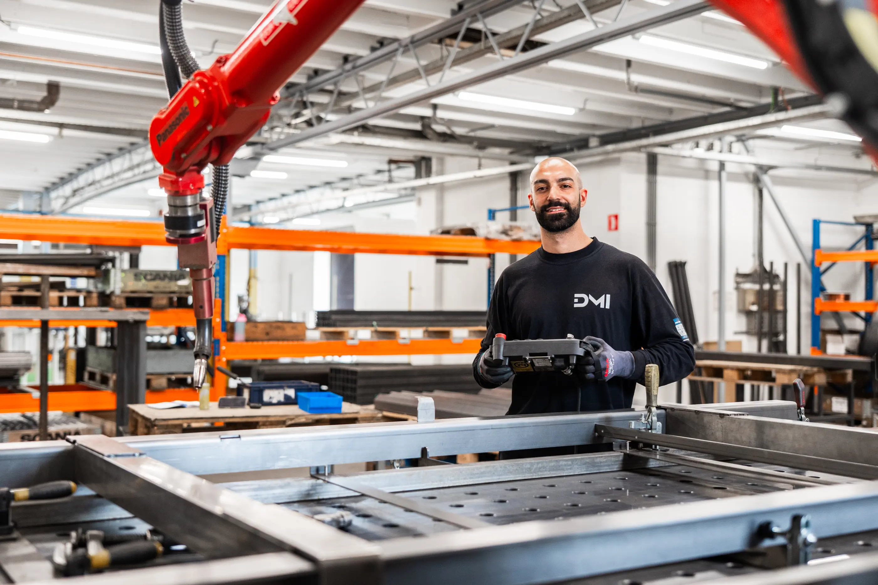 Man met baard en zwart shirt houdt een bedieningskastje vast naast een rode Panasonic-lasrobot in een werkplaats met stellingen.