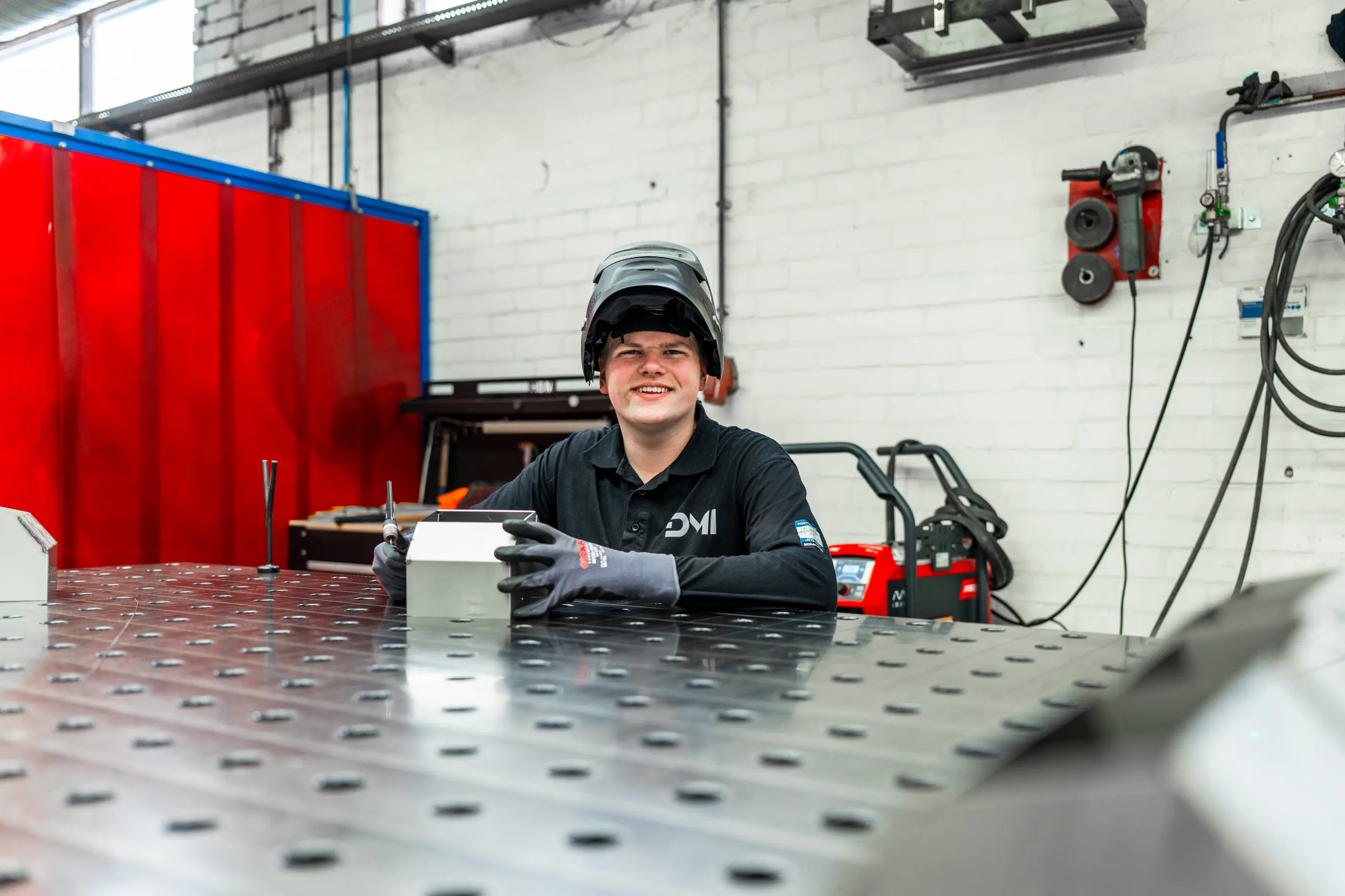 Lachende lasser met opgeklapt lashelm en handschoenen achter een metalen lastafel in een werkplaats met rood scherm.