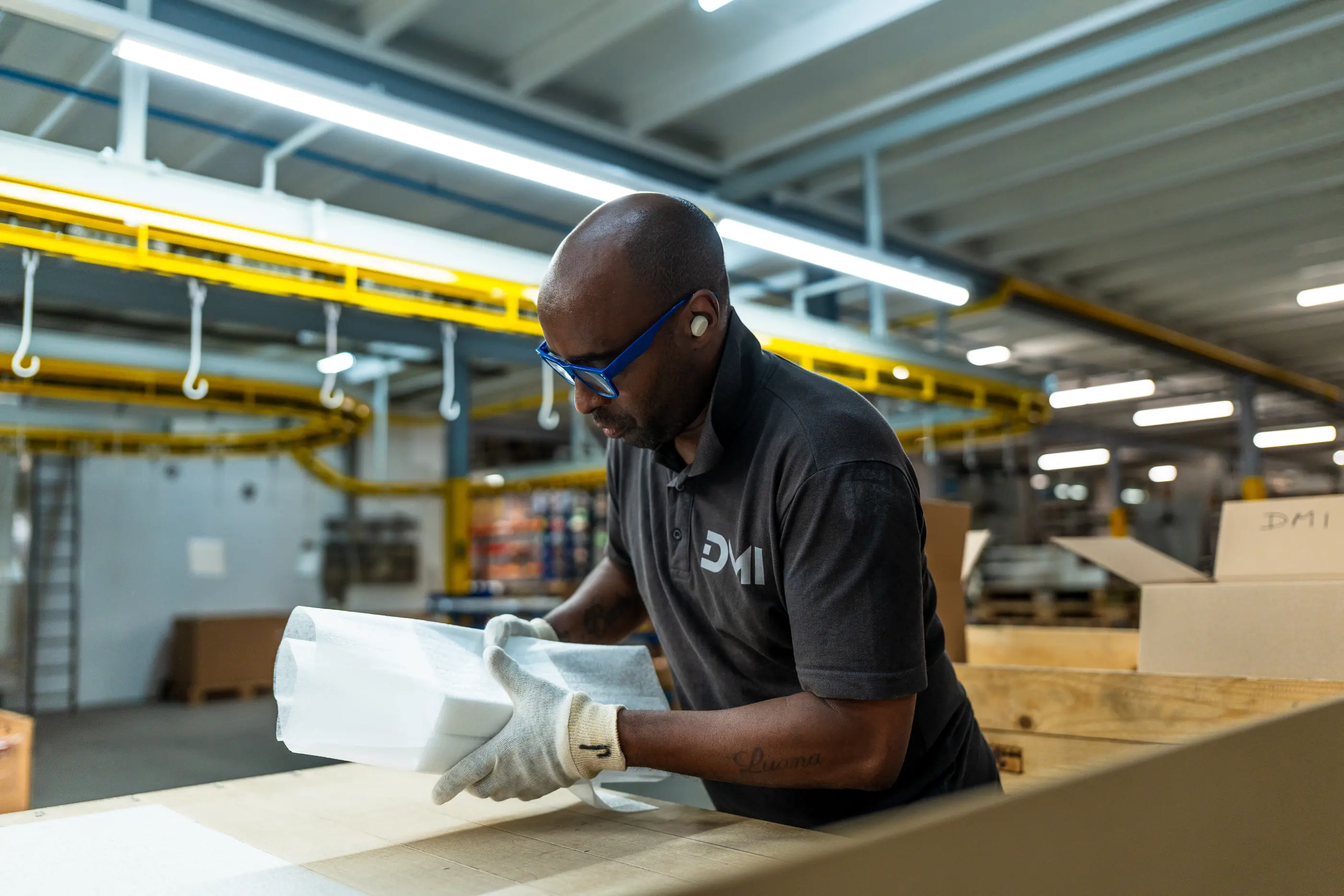 Medewerker in zwart poloshirt met blauwe bril en werkhandschoenen pakt een wit product in witte verpakking in een industriële hal.
