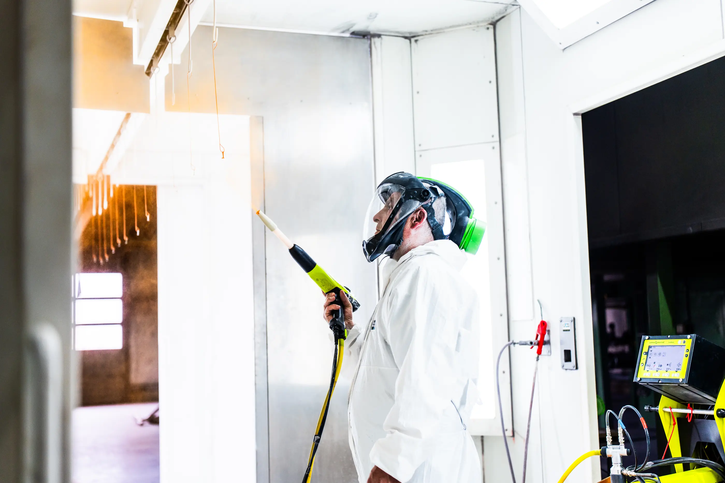 Medewerker in witte overall en gezichtsscherm spuit poedercoating met een spuitpistool in een lichte spuitcabine.