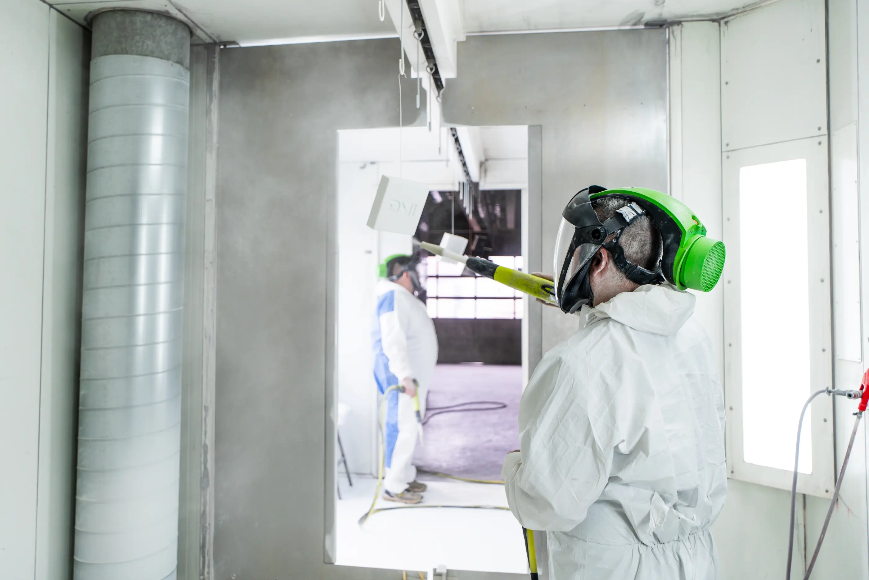 Medewerker in wit beschermend pak met gezichtsscherm en groen filter spuit een werkstuk in een spuitcabine; op de achtergrond een collega.