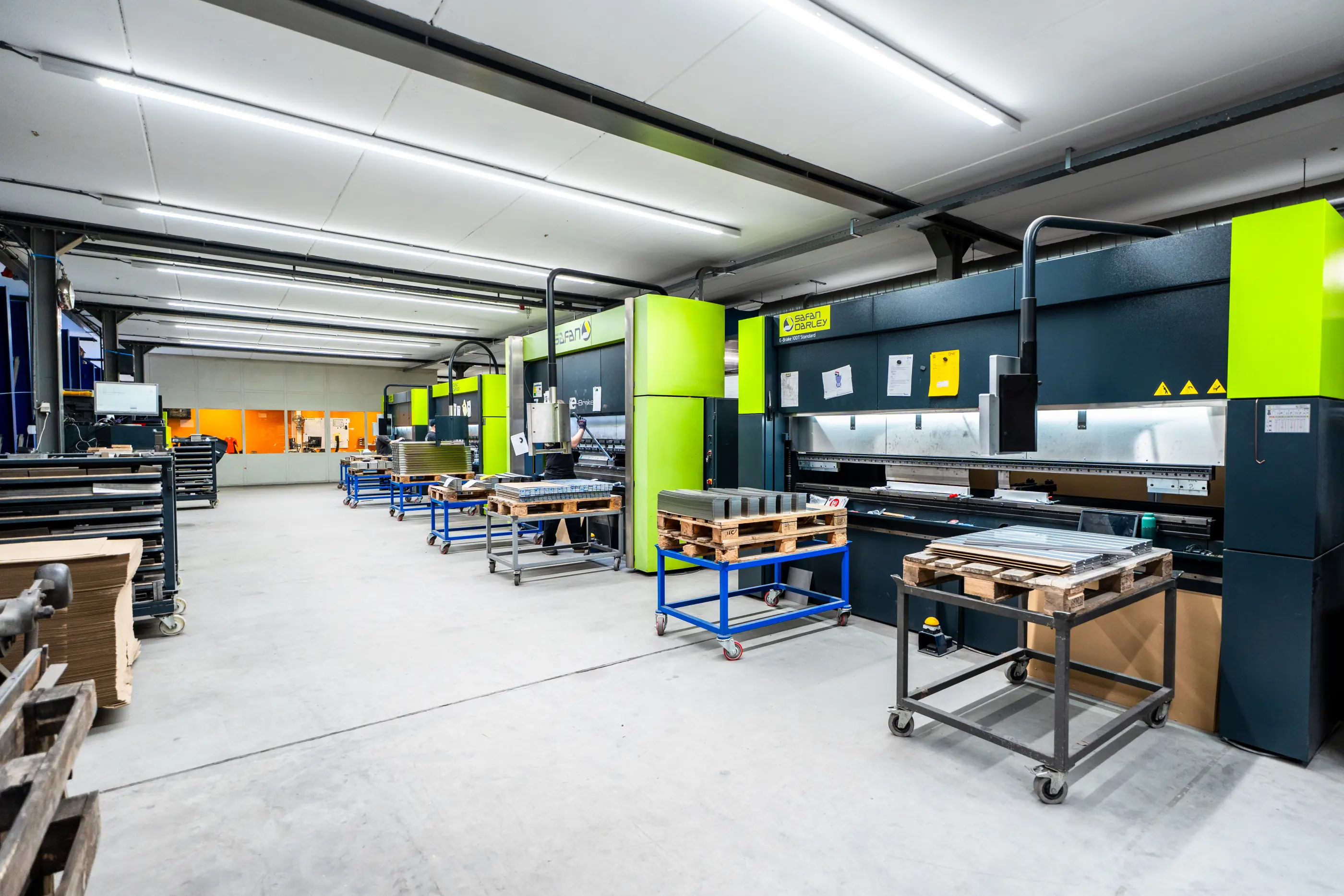 Werkhal met rij groen-zwarte kantbanken en karren met metalen platen op pallets, onder tl-verlichting op een betonnen vloer.