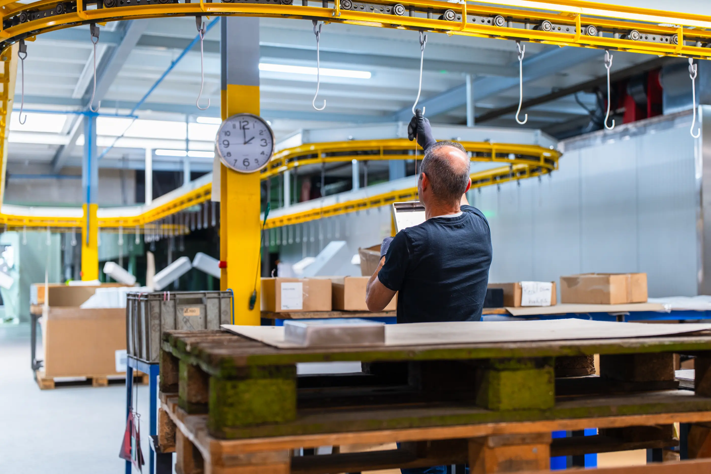 Man in donker T-shirt hangt een metalen plaat aan een haak van een gele transportband in een werkhal met dozen en pallets.