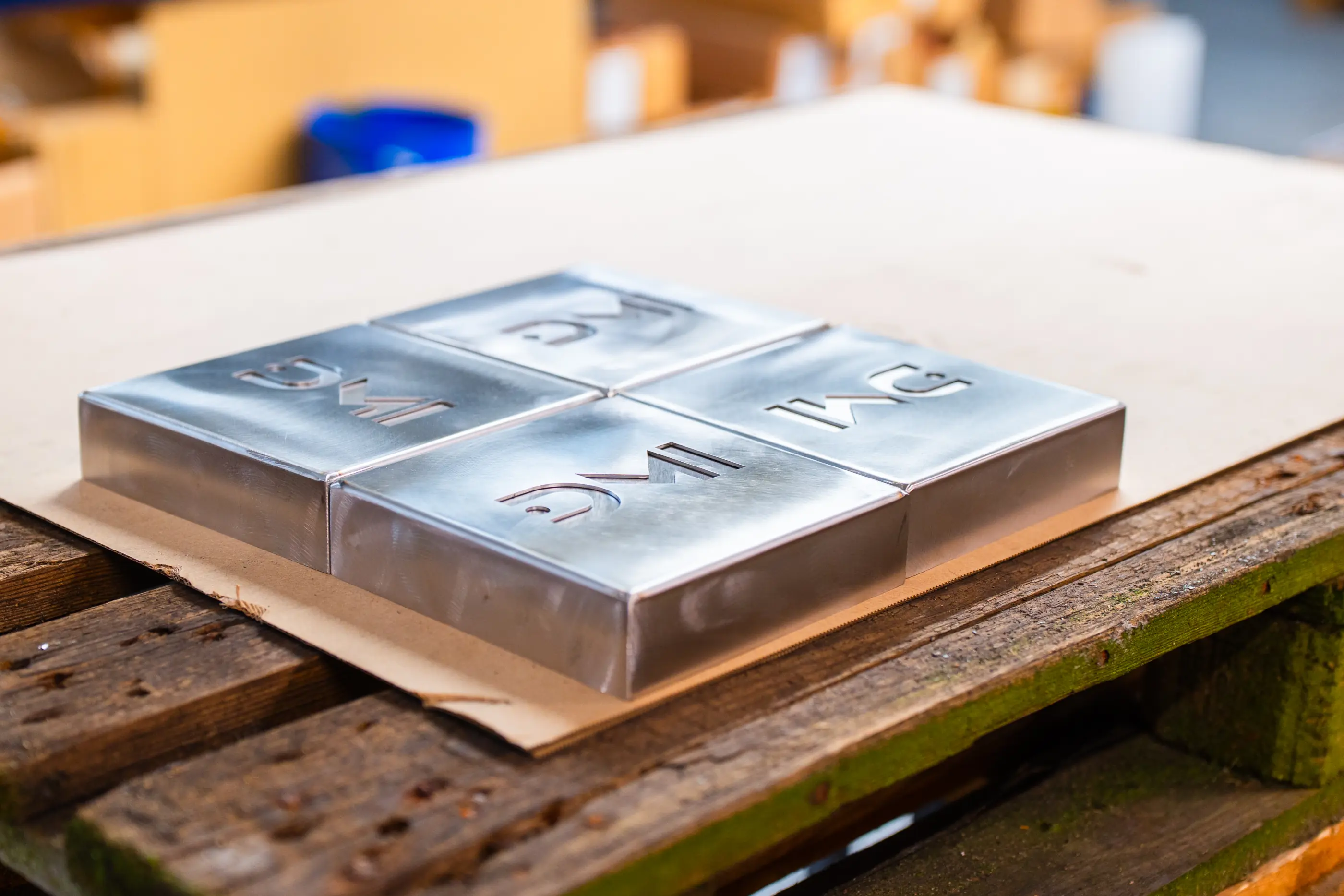 Vier glimmende metalen bakjes met uitgesneden letters liggen op karton op een houten pallet in een werkplaats.