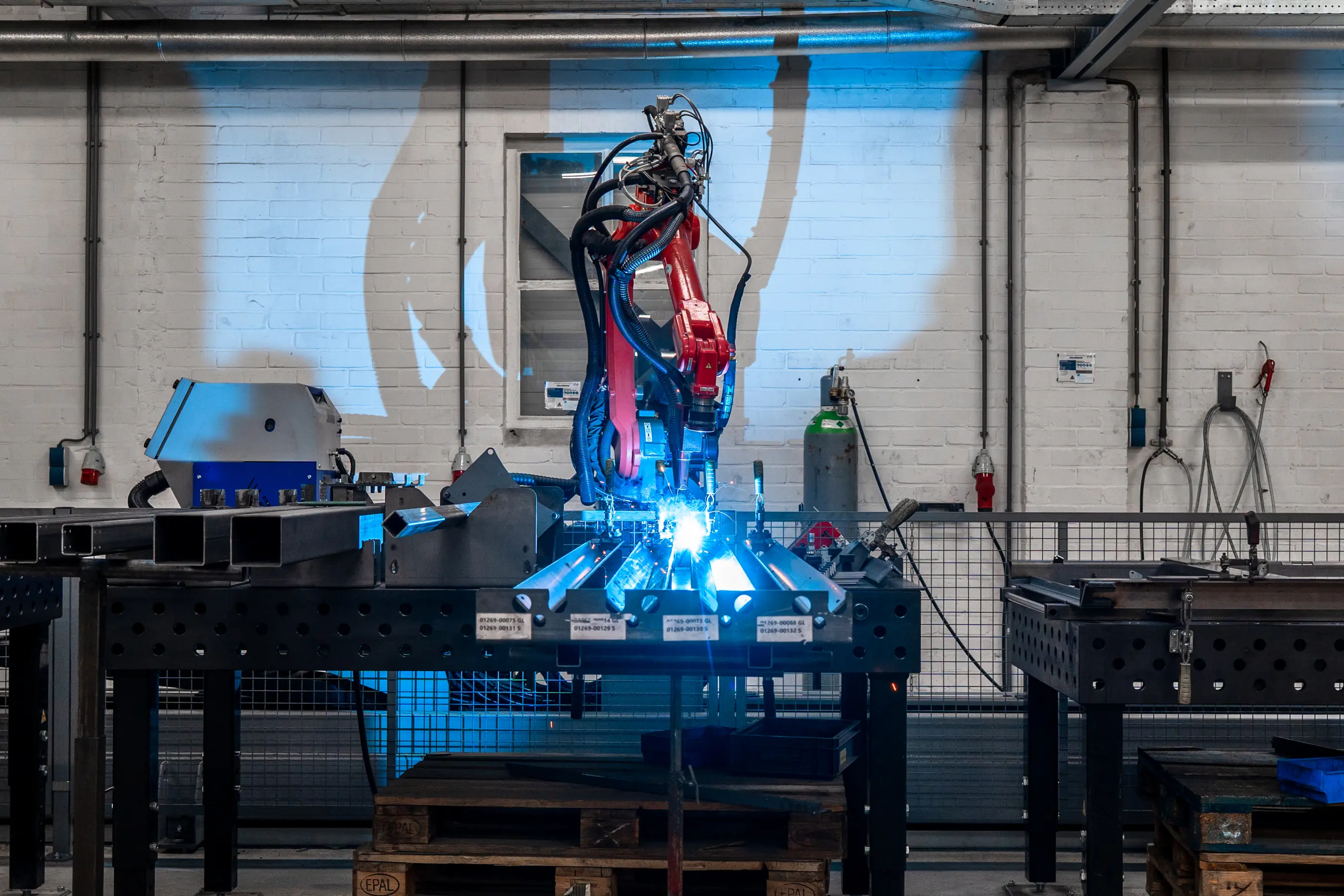 Rode lasrobot last stalen profielen op een werktafel in een werkplaats met witte bakstenen muur en blauw licht.