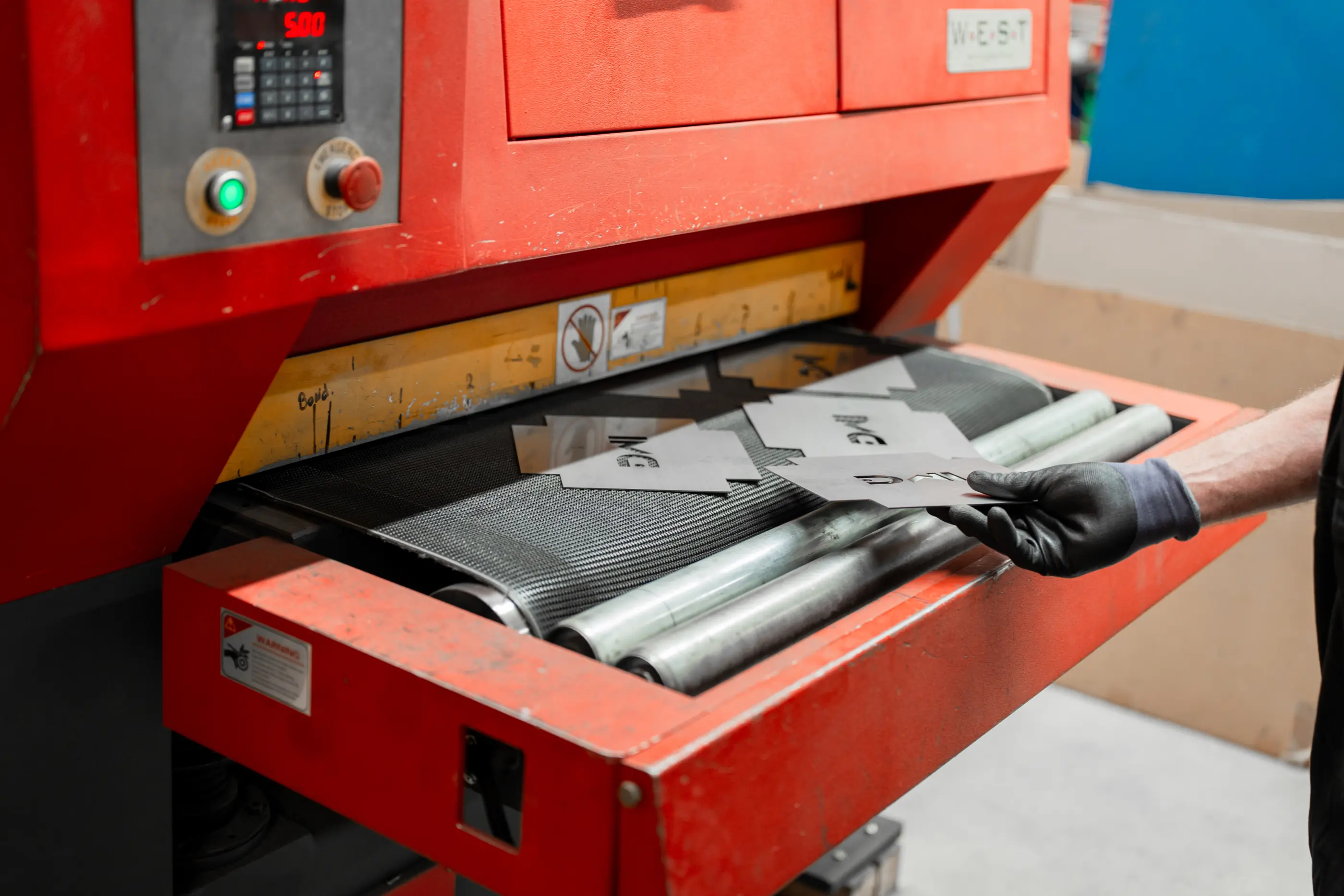 Hand met zwarte werkhandschoen legt metalen plaatjes op de transportband van een rode machine met digitaal display.